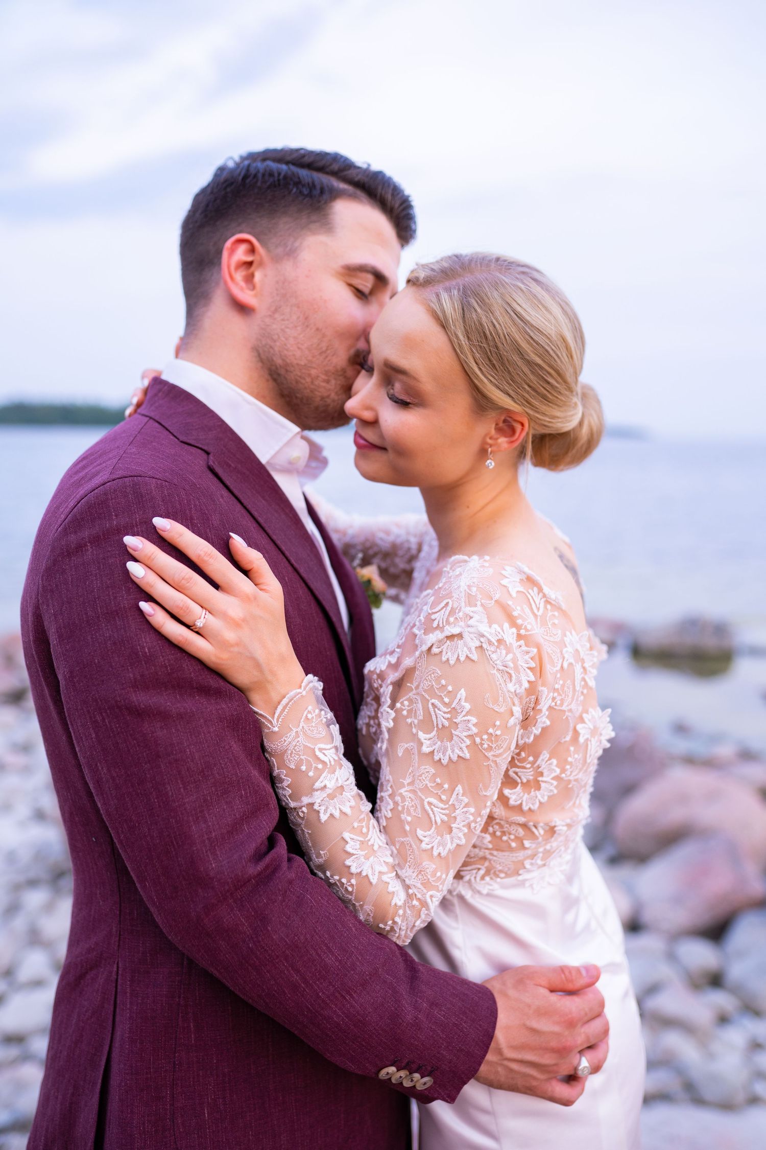 Bride and groom in tender embrace on rocky beach, bride in lace gown and groom in burgundy suit at twilight.