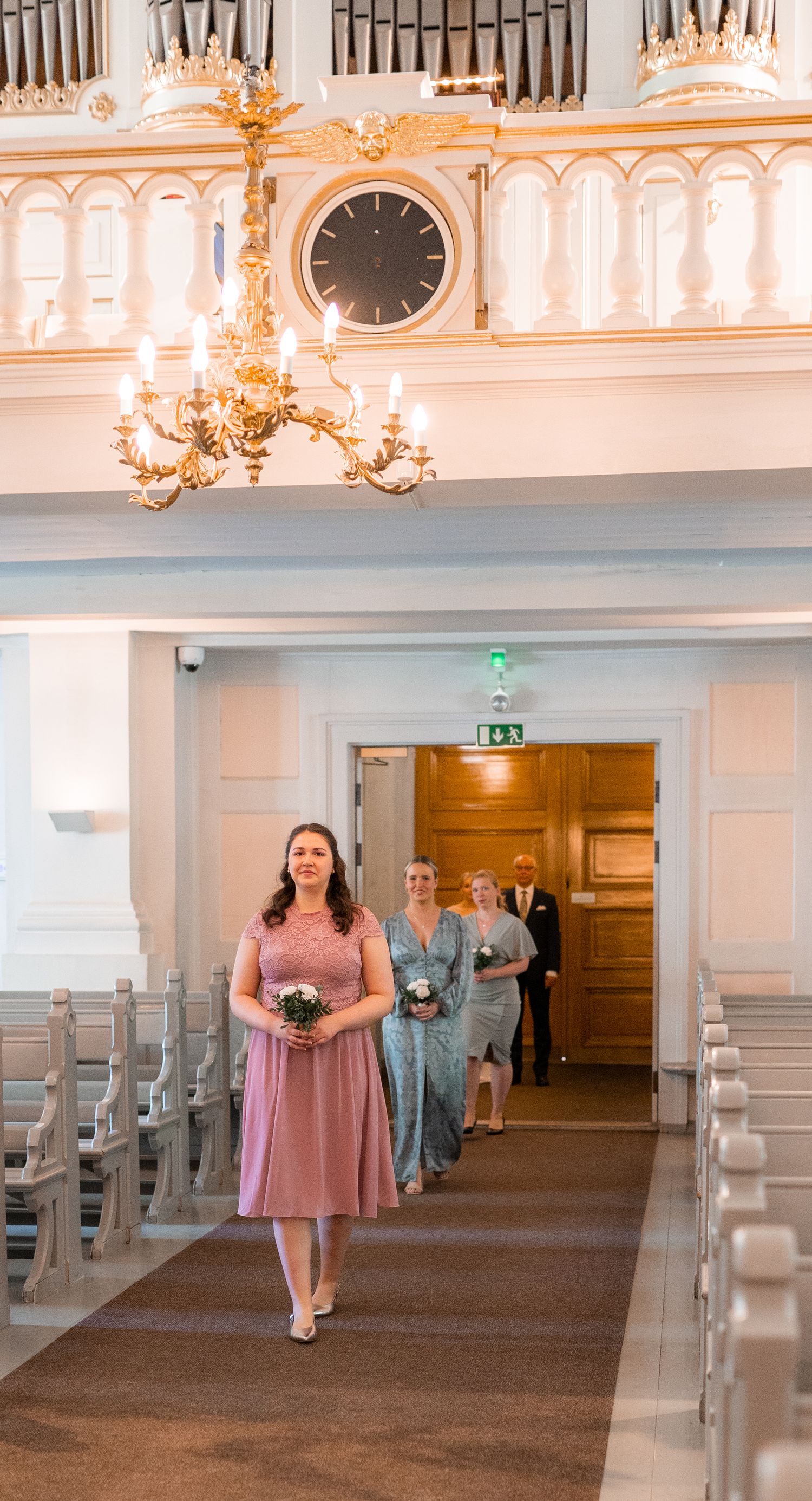 Bridesmaids in pink and blue dresses walking down elegant white church aisle with bouquets.