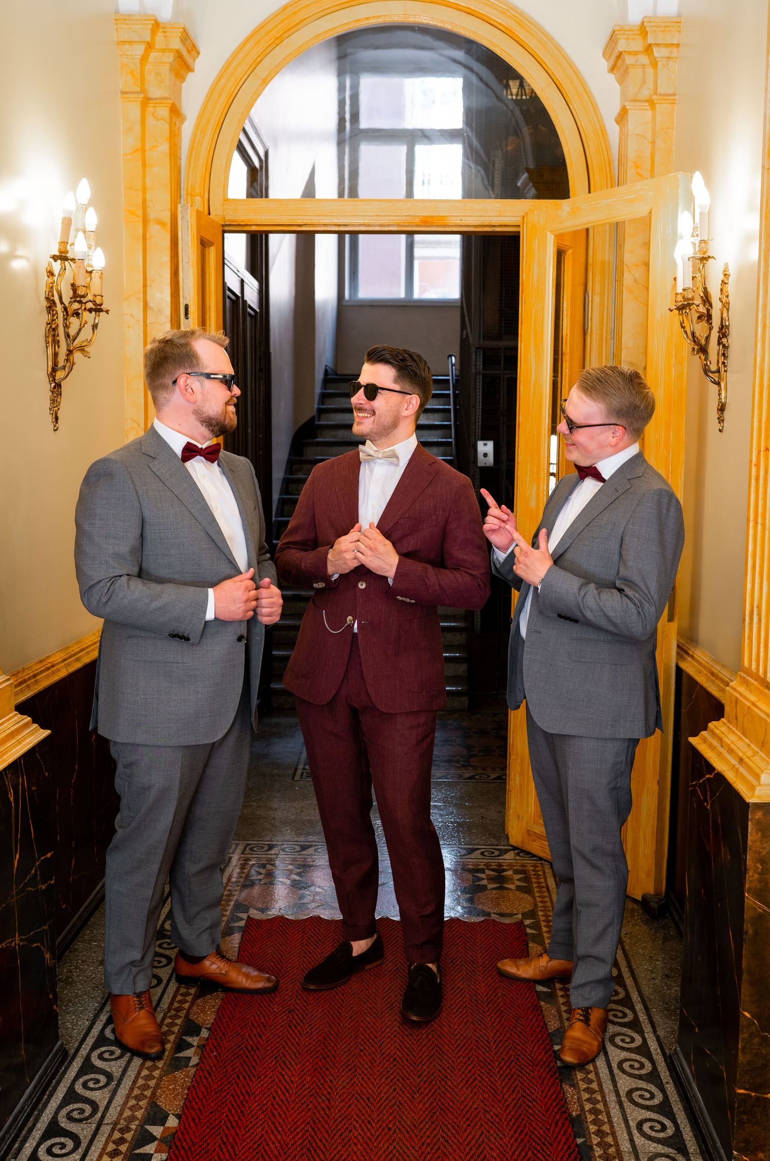 Groom in burgundy suit with groomsmen in grey suits laughing together in a grand hallway.