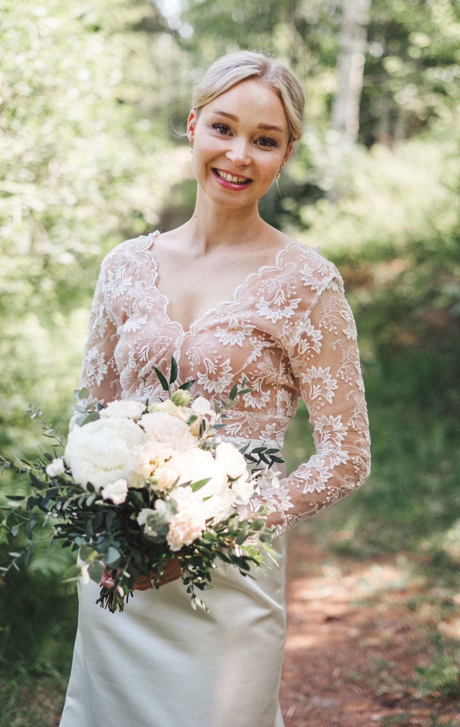 Smiling bride in lace long-sleeve wedding gown holding white peony bouquet outdoors.