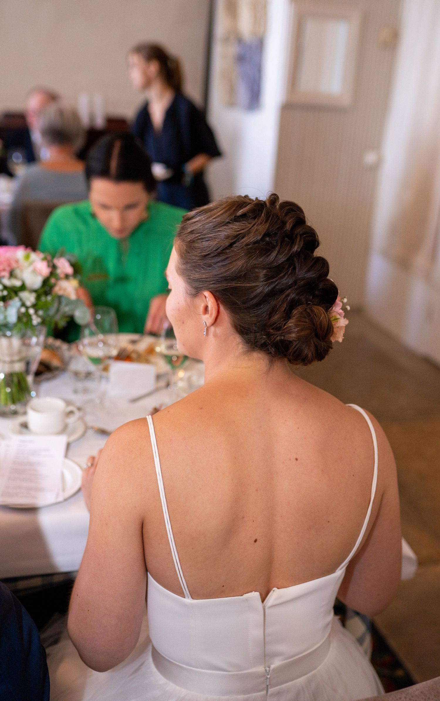 Bride in white spaghetti-strap gown sits at wedding reception table showing elegant braided updo hairstyle.