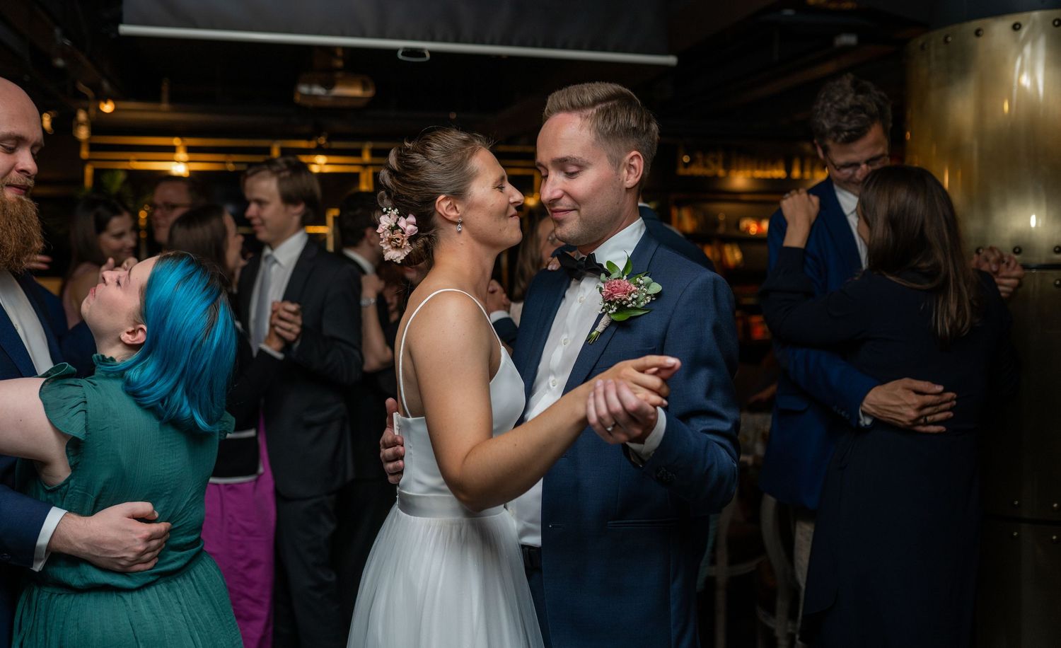 Bride in white dress and groom in blue suit share a romantic dance surrounded by celebrating wedding guests.