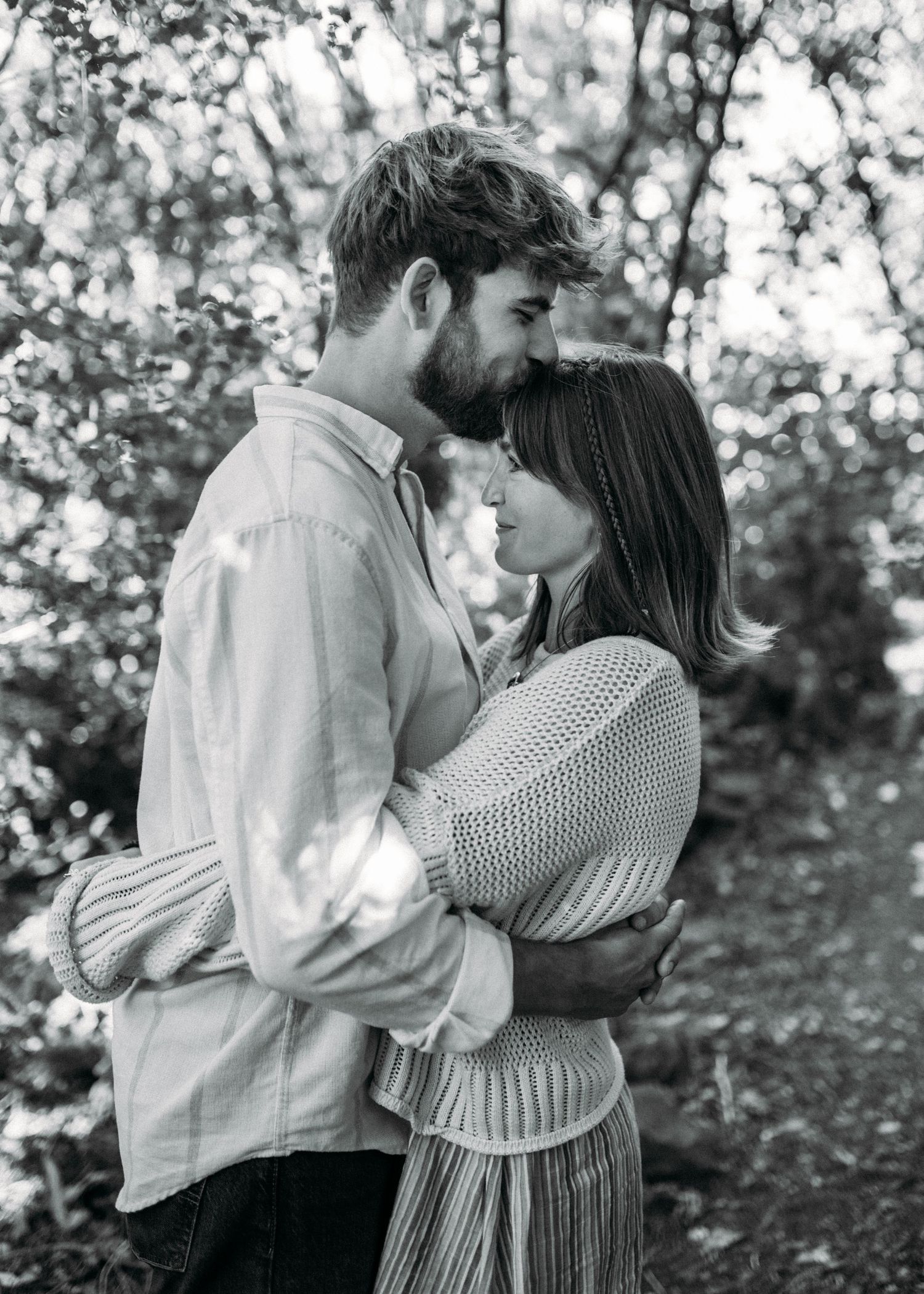 Black and white photo of couple sharing an intimate moment embracing in a forest path.