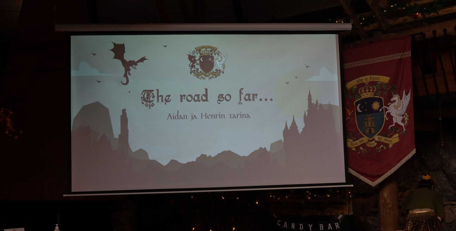 Presentation slide reading 'The road so far' with medieval fantasy artwork displayed in a darkened conference room.