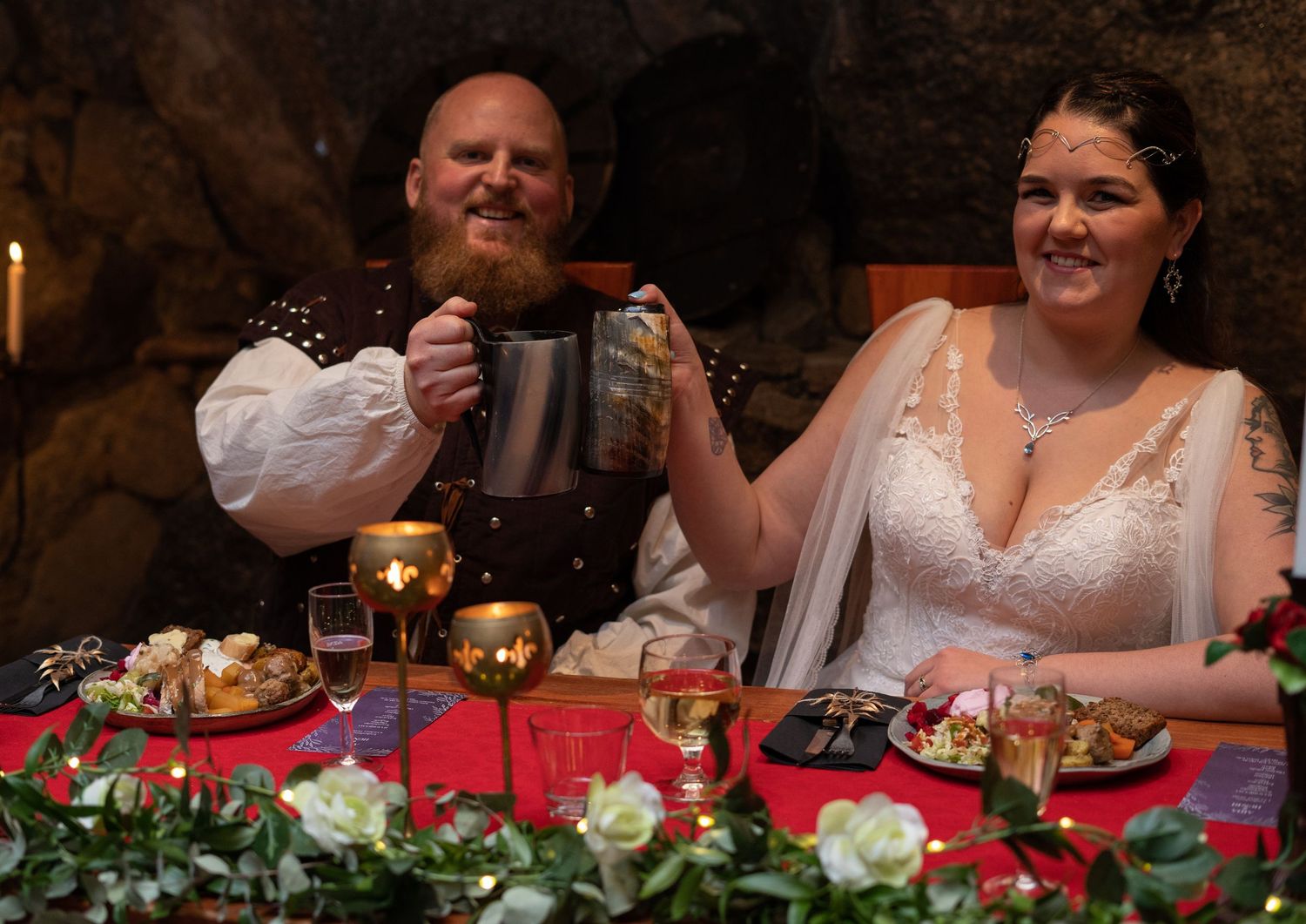 Medieval wedding couple toasting with metal tankards at a decorated banquet table in a stone castle setting.