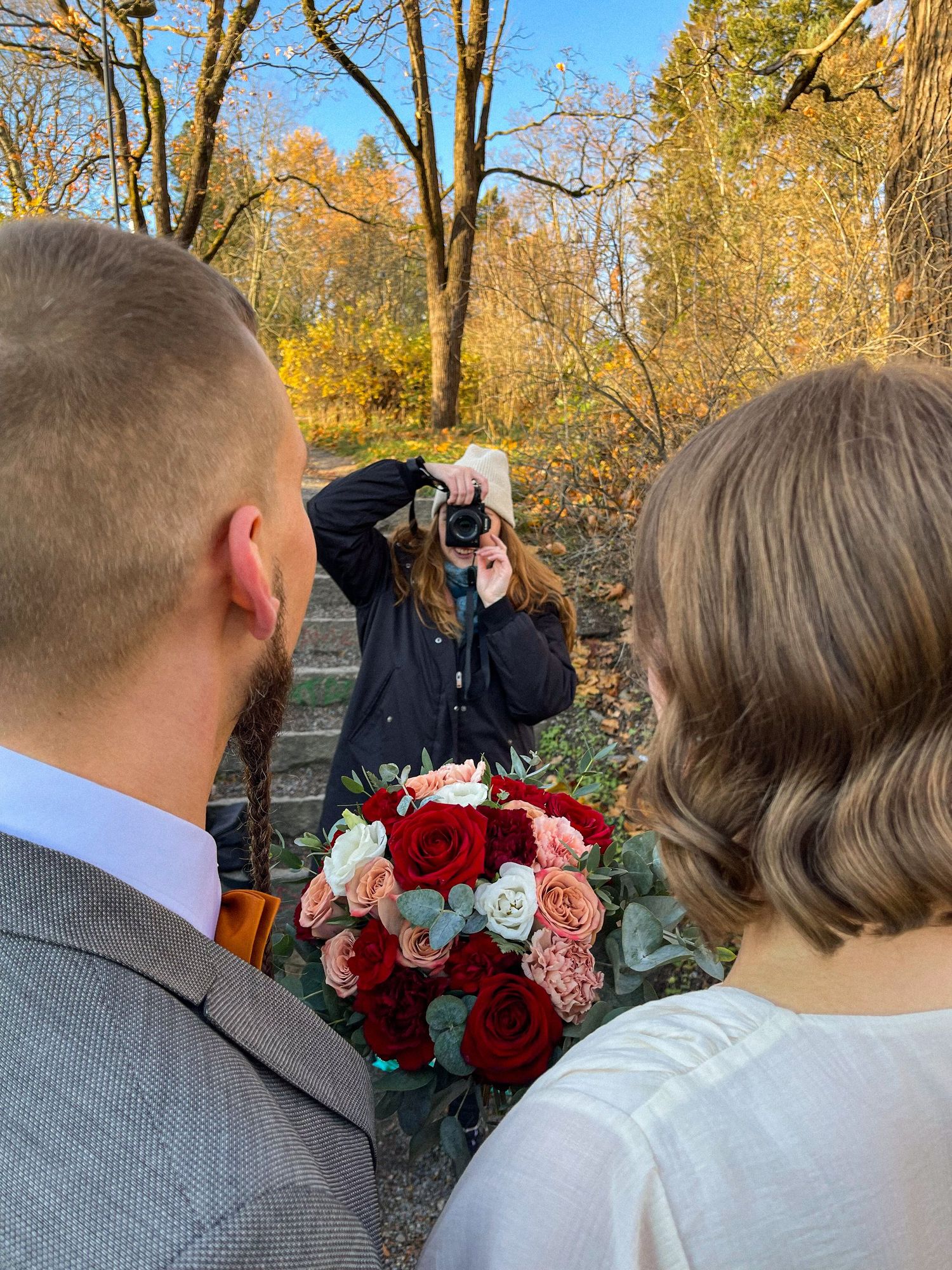 Photographer taking pictures of a couple holding a red and pink rose bouquet in a scenic autumn outdoor setting.