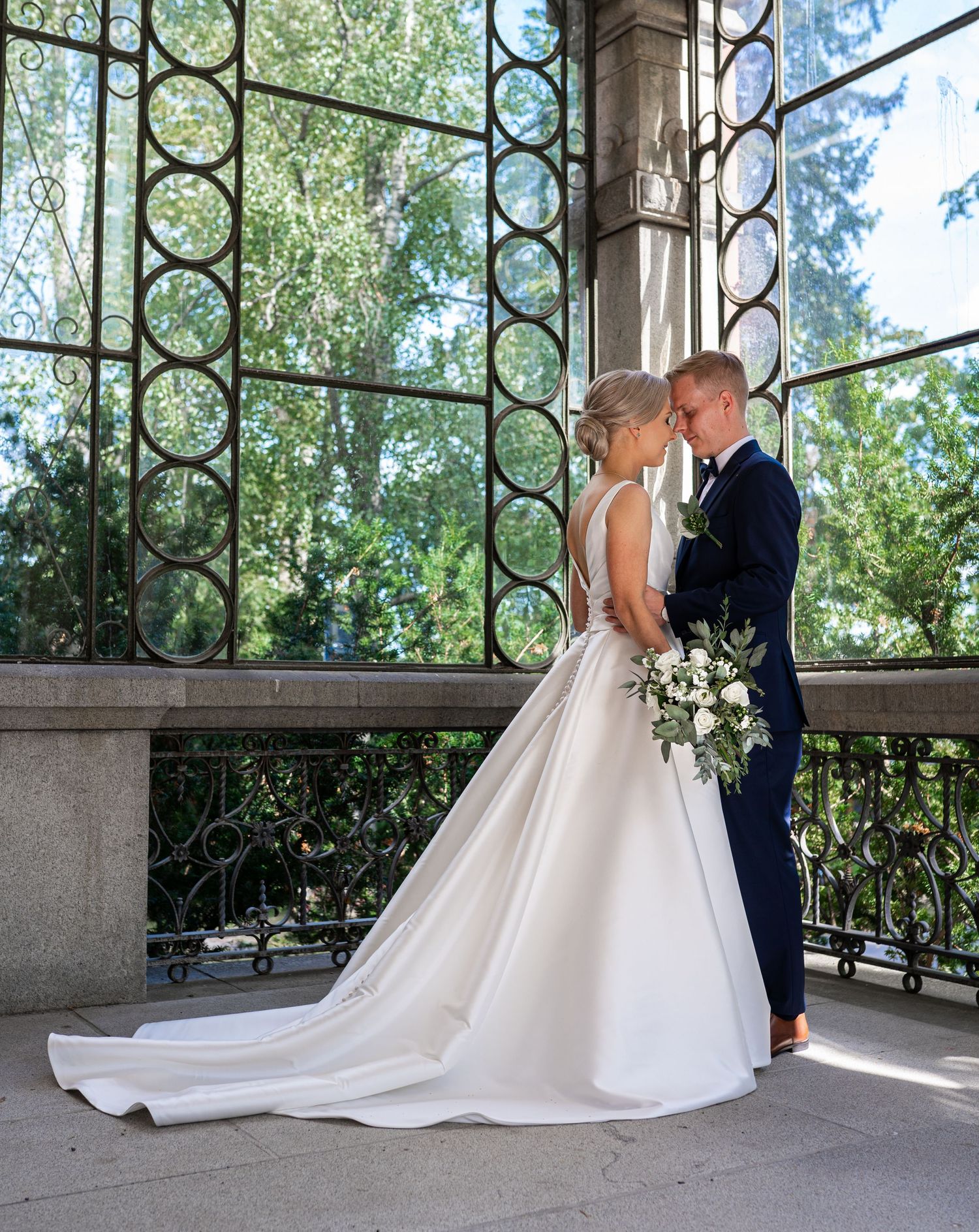 Wedding couple embrace tenderly on a stone balcony with ornate ironwork and lush green trees.