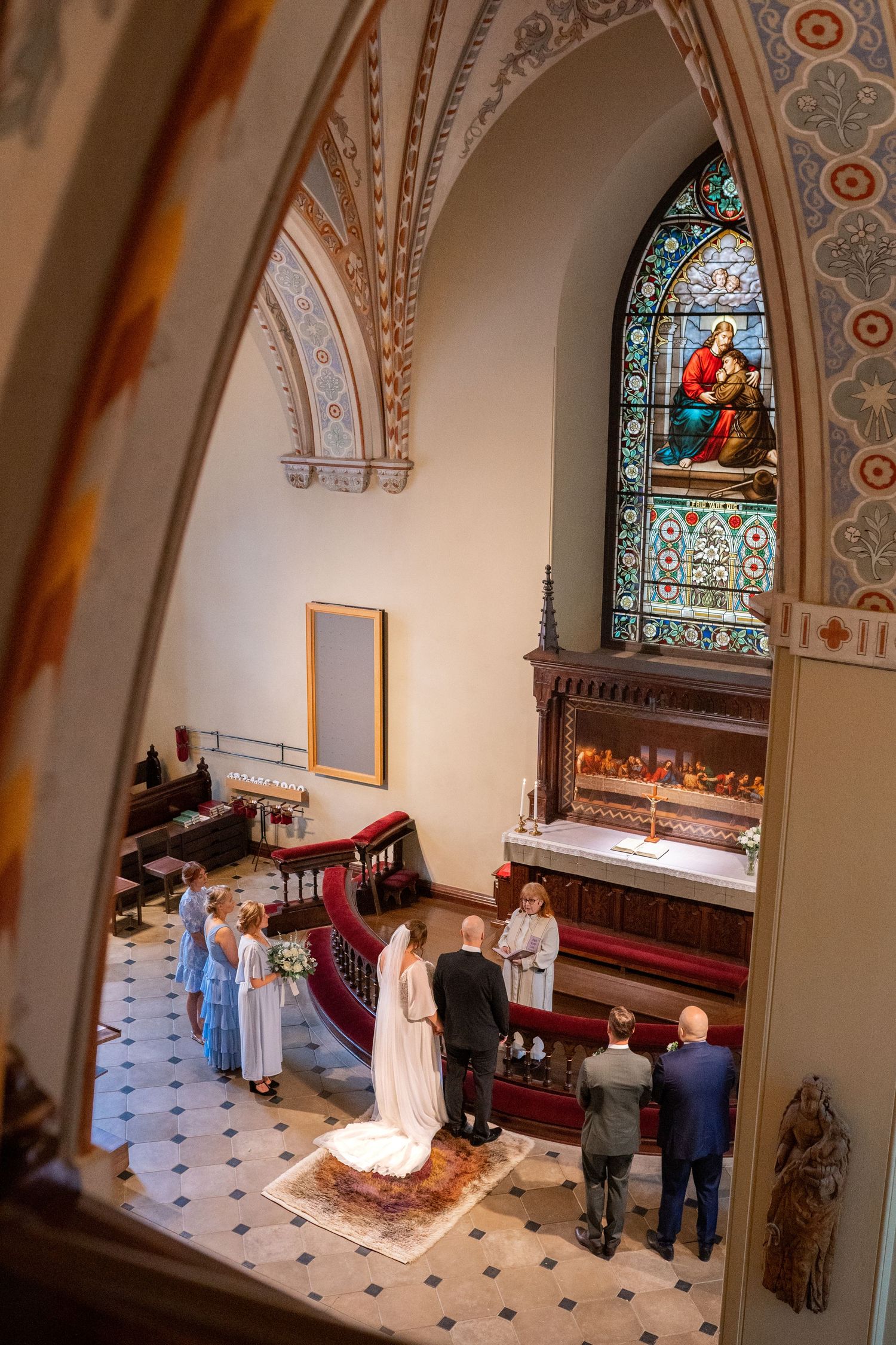 Aerial view of wedding ceremony at ornate church altar with stained glass window and bridal party.