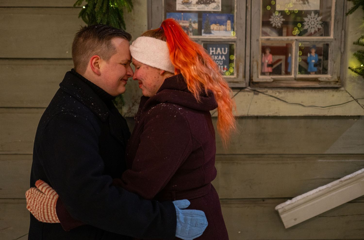 Couple embracing outdoors at night, woman with orange hair wearing pink hat, man in black jacket, near decorated window.