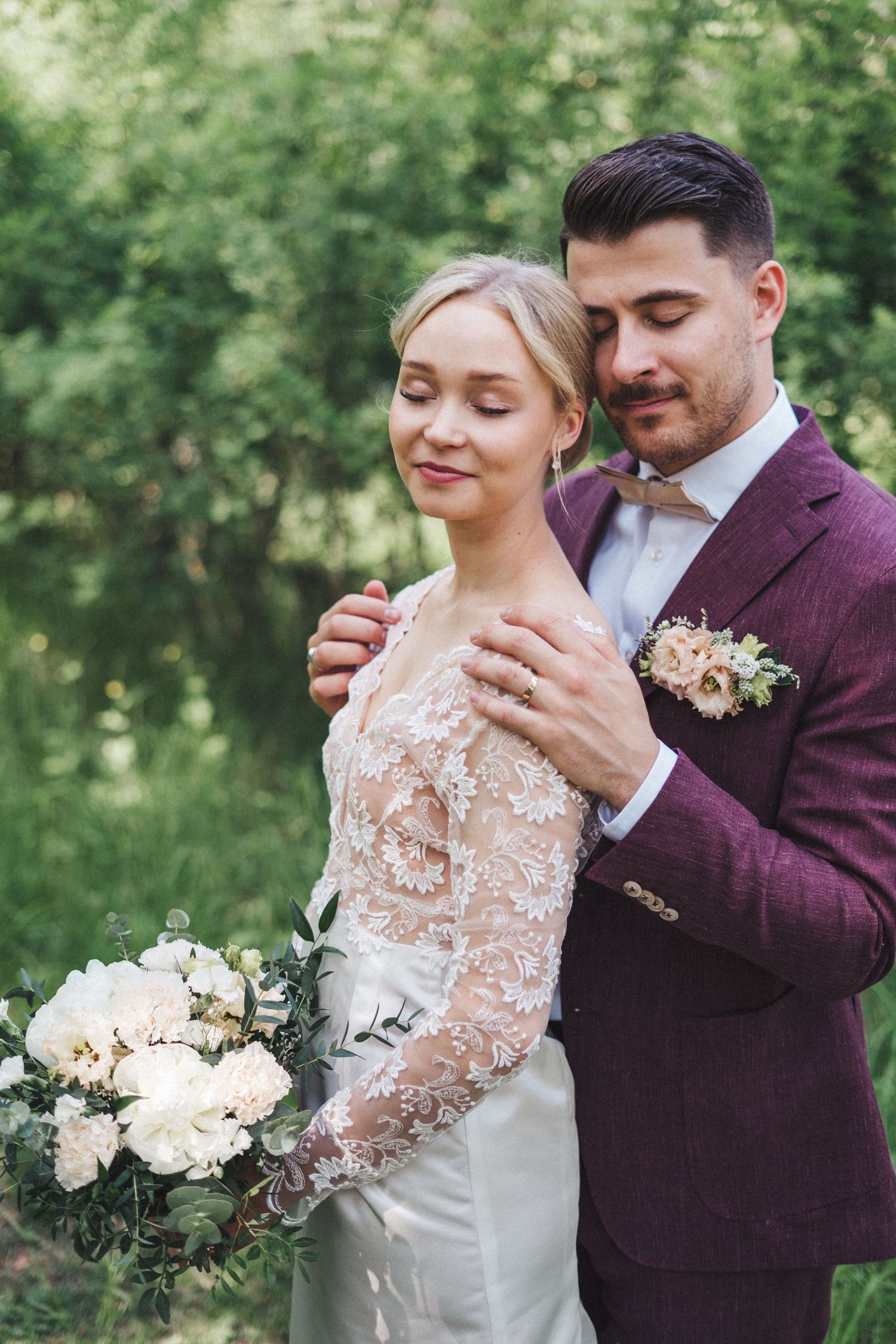 Groom in burgundy suit tenderly embracing bride in lace gown with eyes closed in a lush green garden.