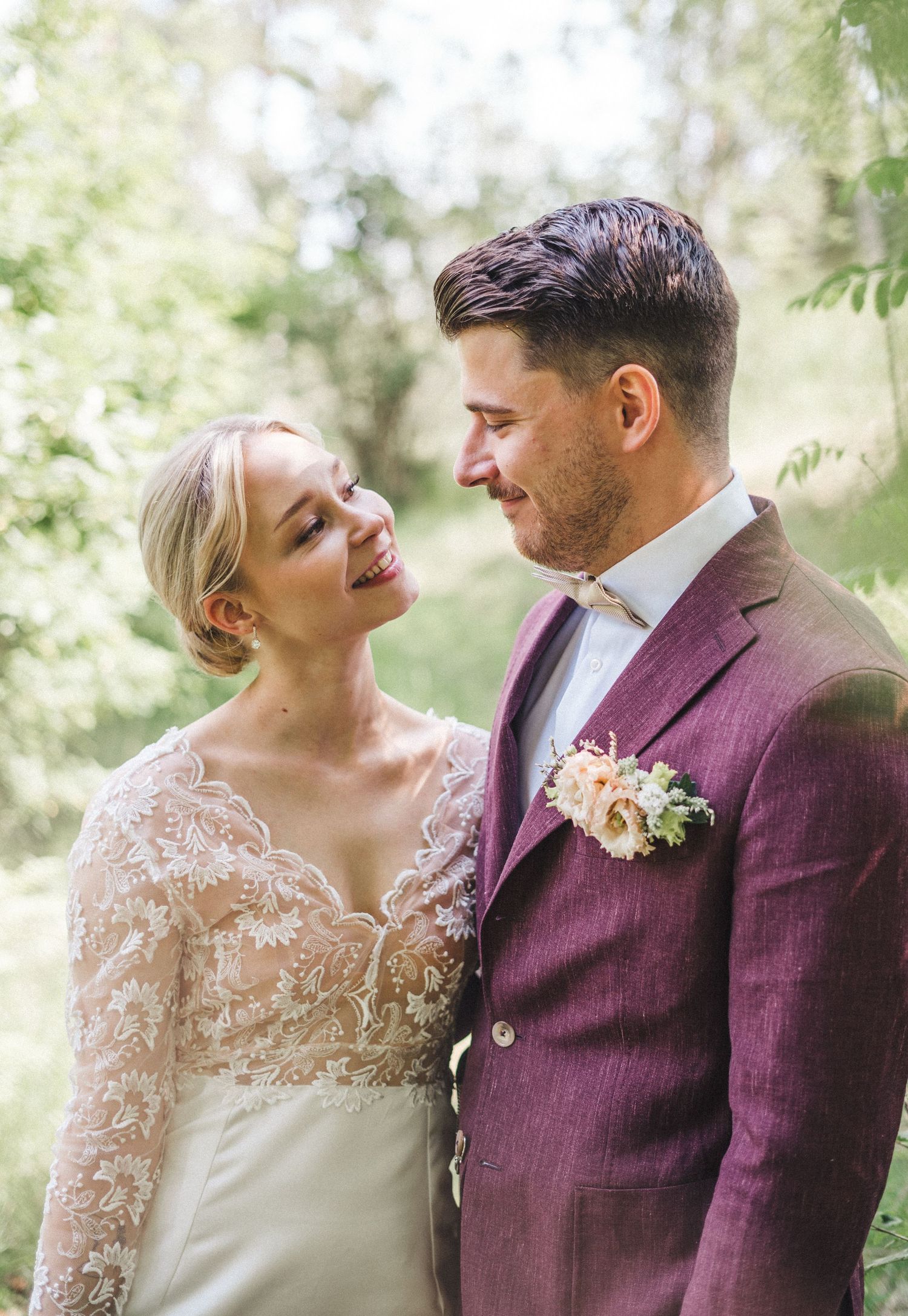 Bride in lace gown and groom in burgundy suit smiling at each other in a sunlit garden setting.