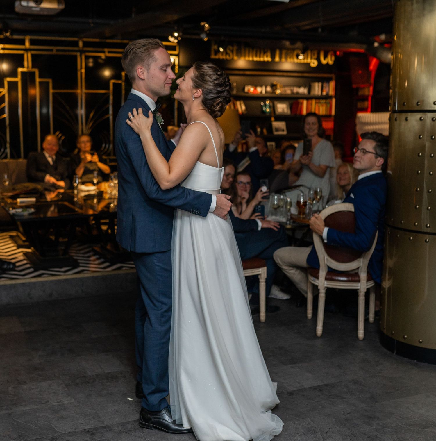 Bride and groom share their first dance at wedding reception while guests watch and applaud in stylish venue.