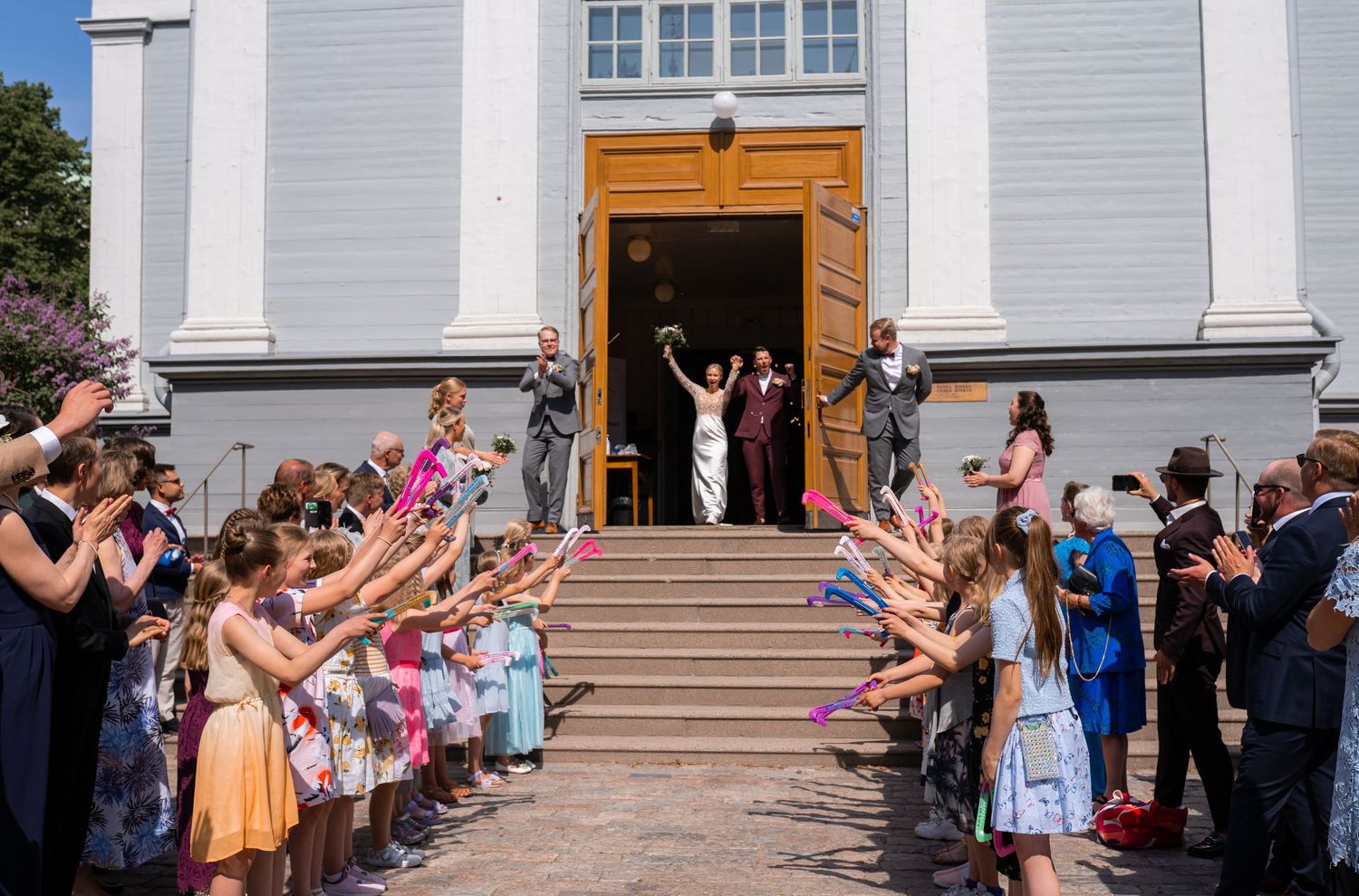 Bride and groom exit church doors to cheering guests waving colorful ribbons on sunny outdoor steps.