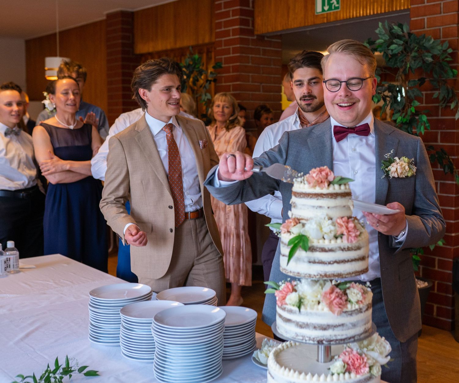 Groom cutting a three-tier naked wedding cake decorated with pink flowers at a wedding reception.