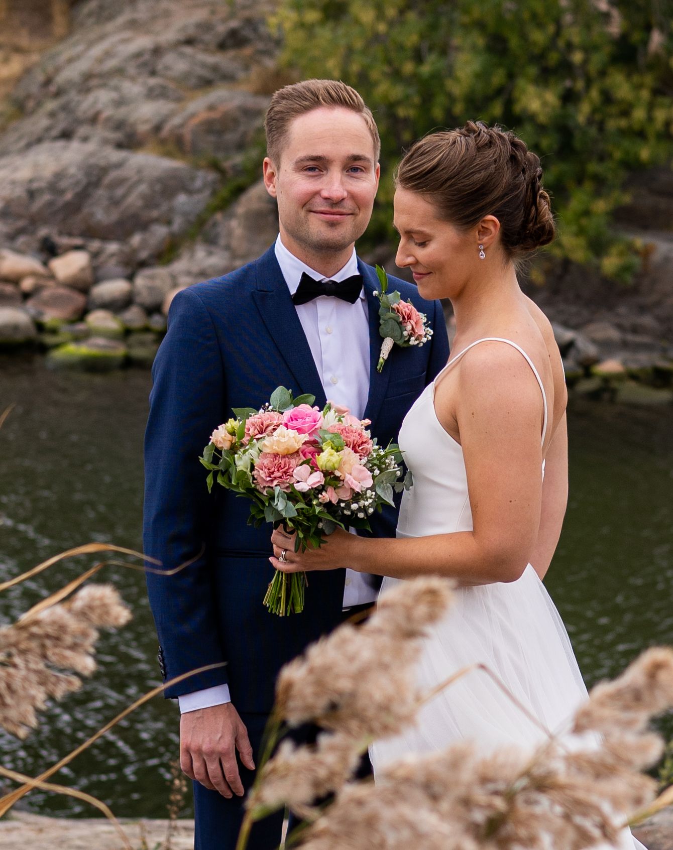 Close-up of groom in navy tux smiling as bride looks down at her pink and green bridal bouquet.