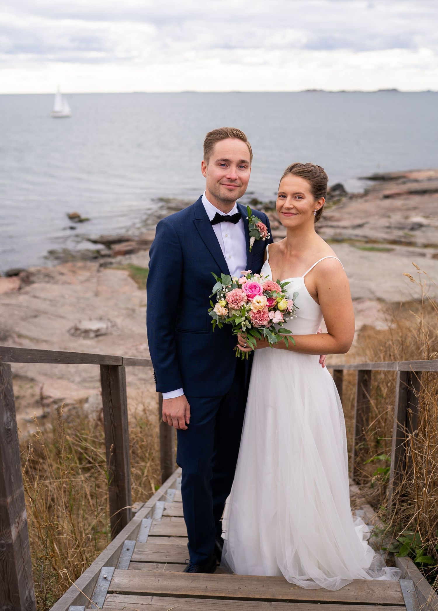 Bride and groom posing on wooden steps by rocky seaside, she holds pink bouquet, he wears navy suit with bow tie.