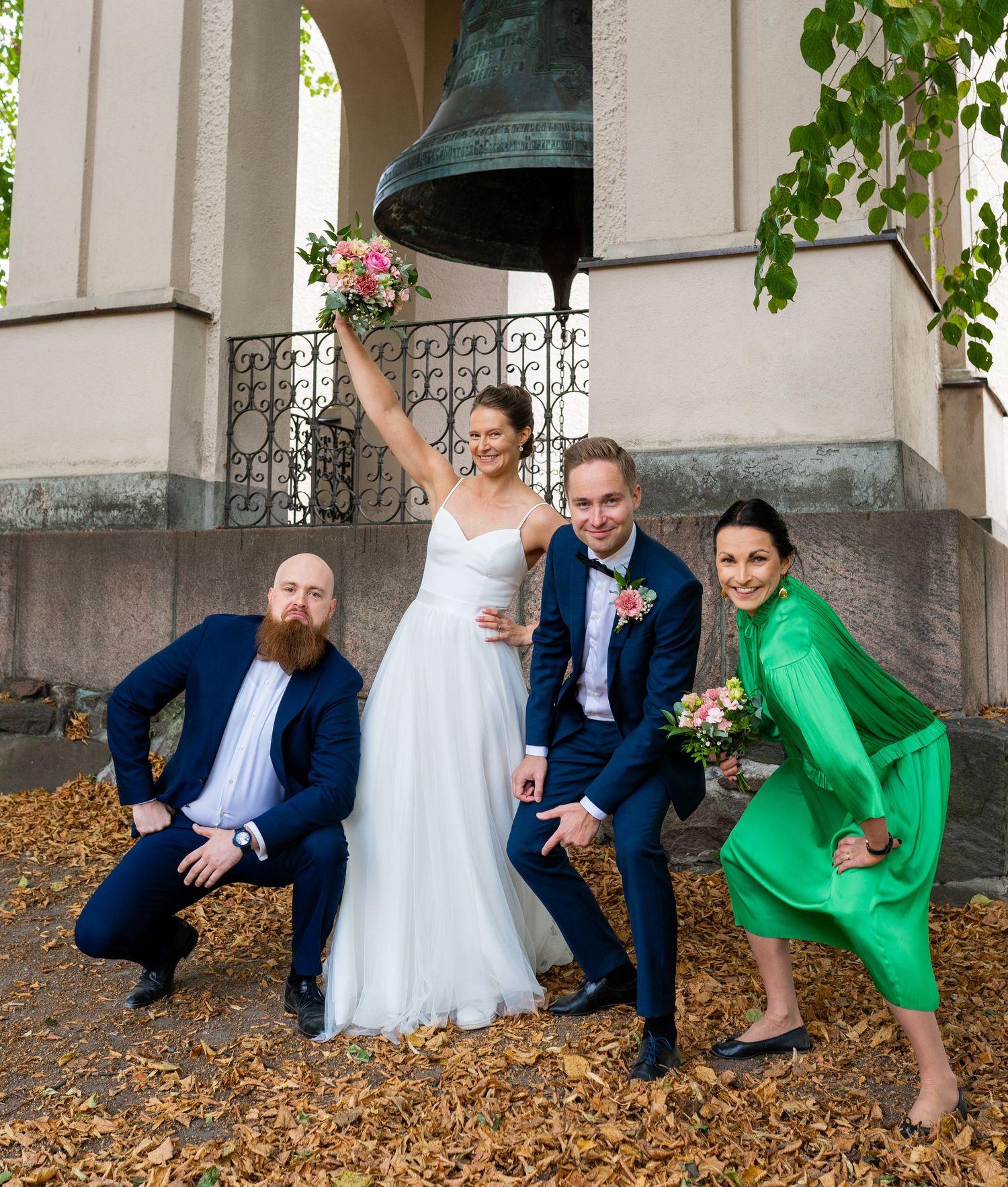 Wedding party of four posing playfully near large church bell surrounded by golden autumn leaves.