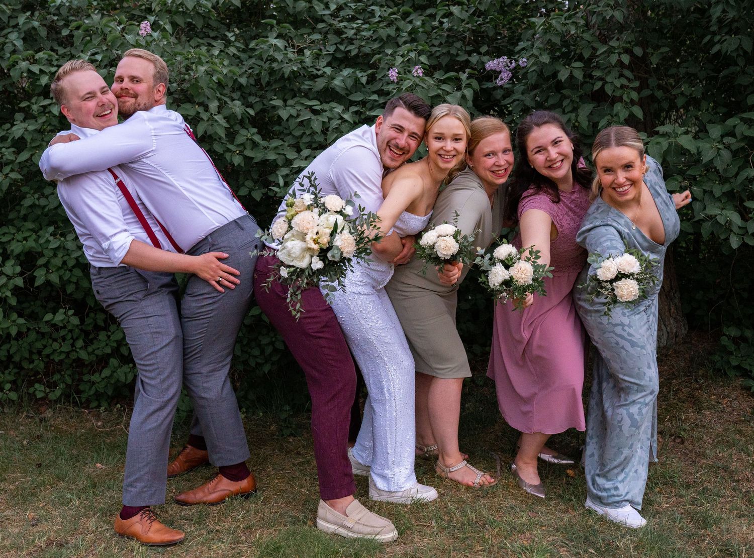 Joyful wedding party hugging and laughing outdoors in burgundy and pink formal attire with bouquets.