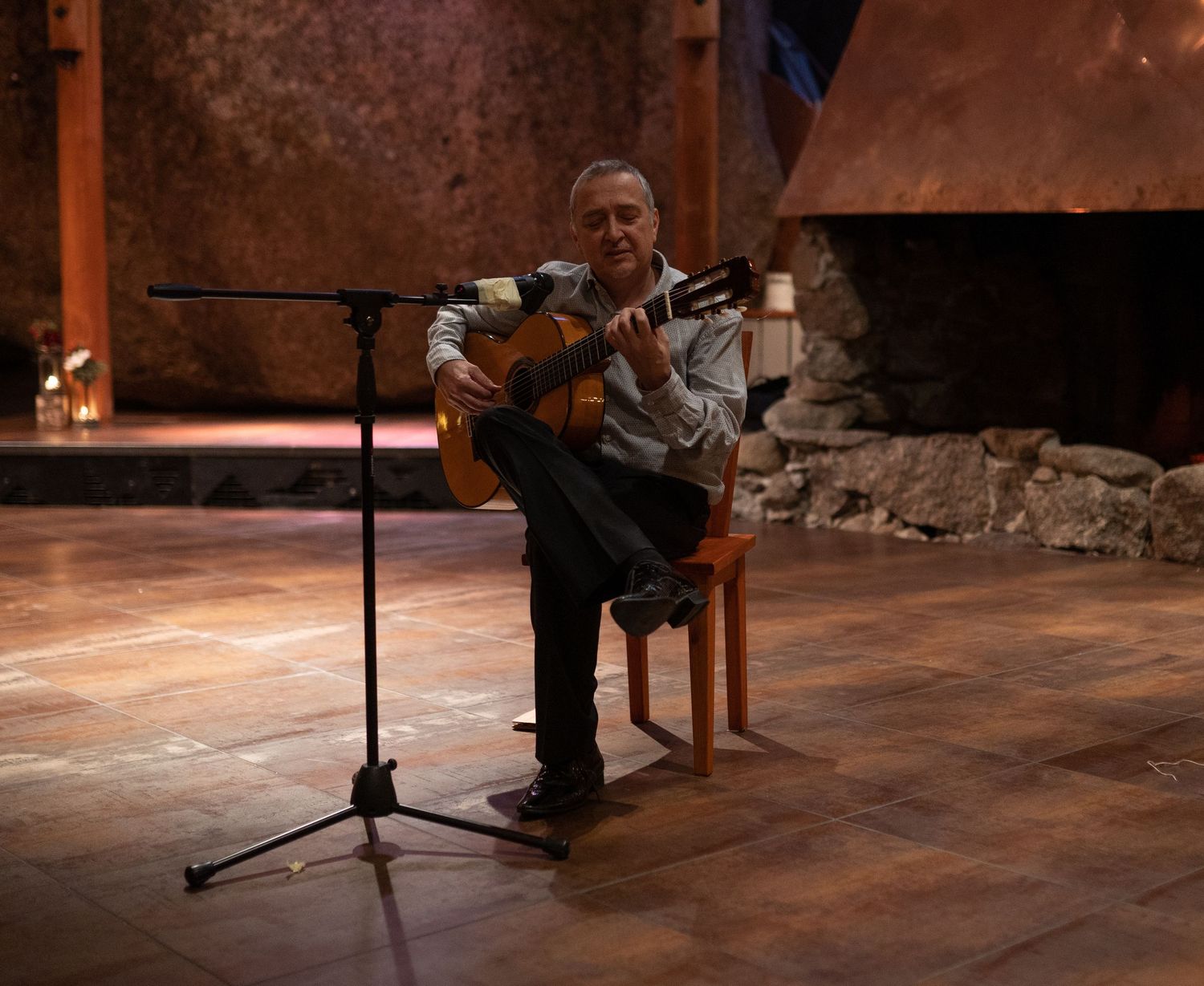 Musician playing acoustic guitar while seated on a stool in front of a microphone stand indoors.
