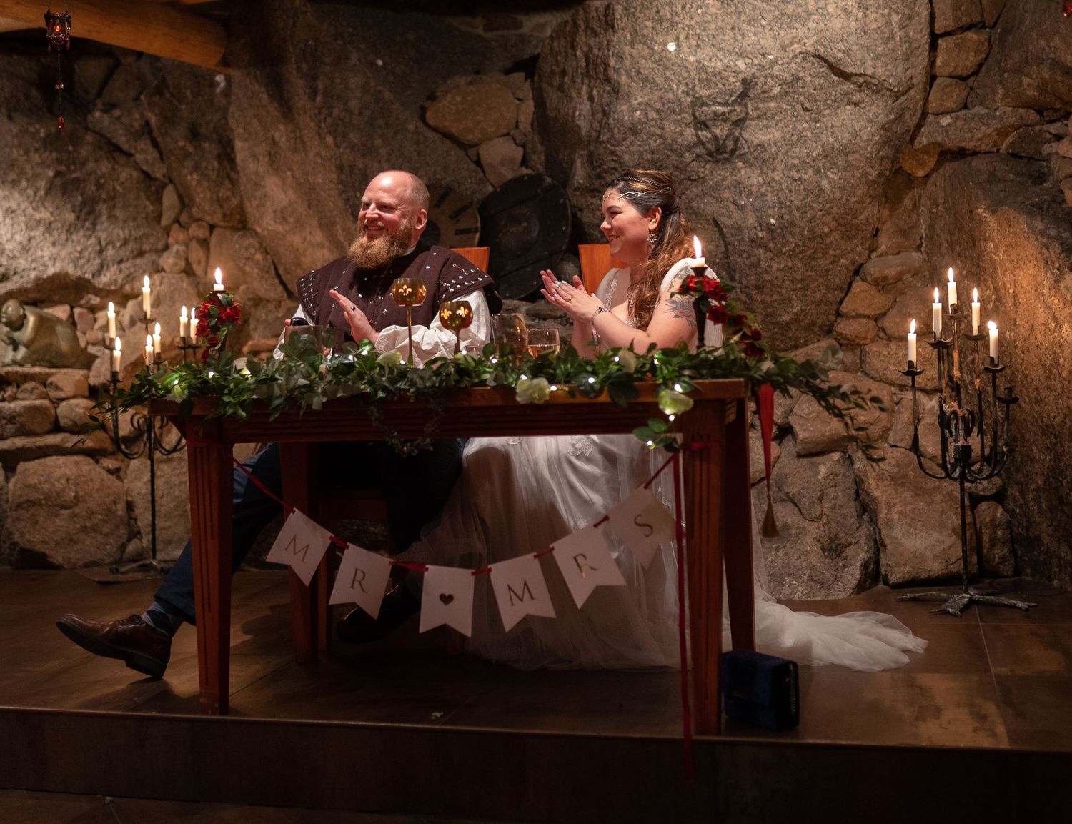 Bride and groom laughing at candlelit sweetheart table with floral garland in rustic stone venue.