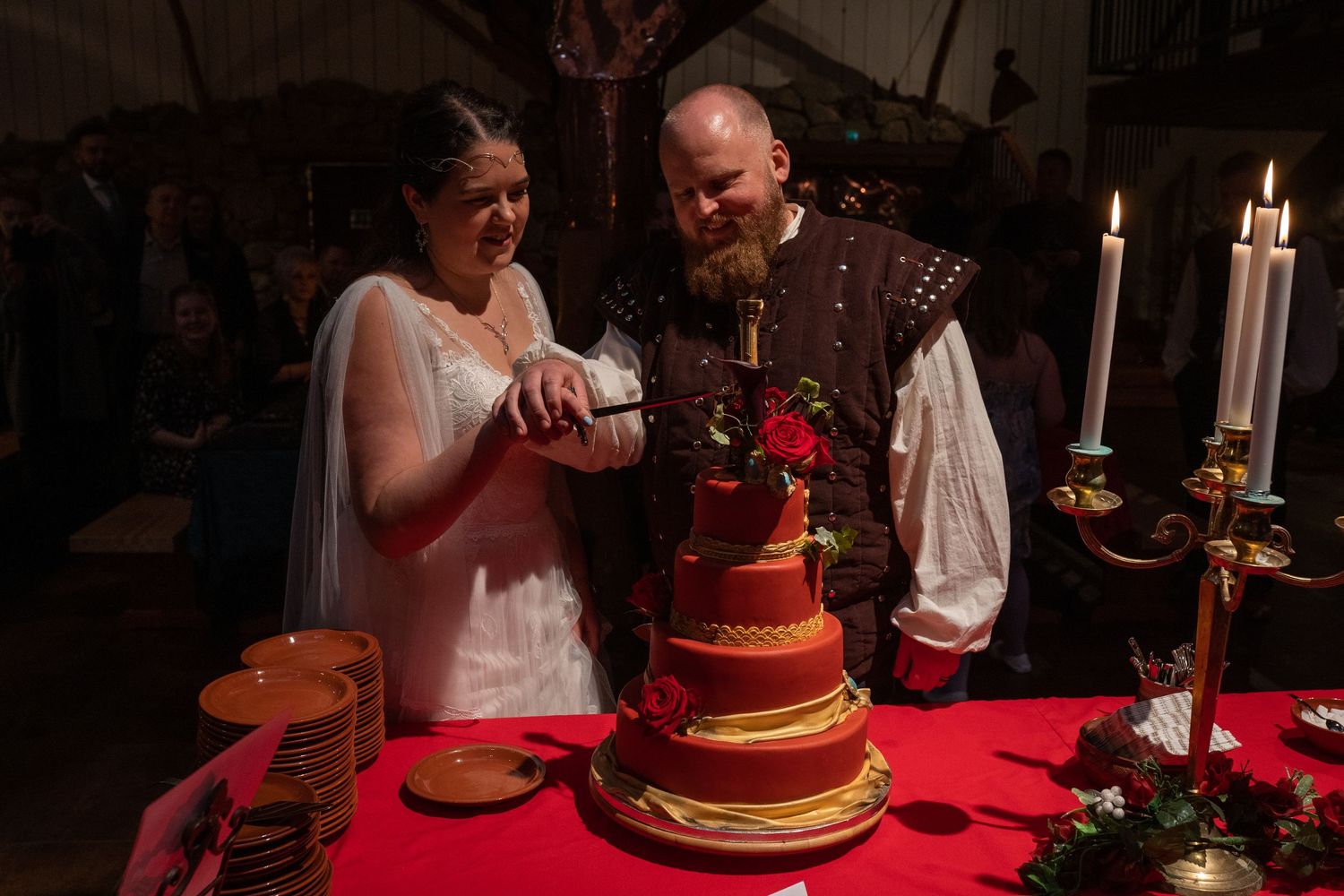 Couple in medieval attire cutting a red tiered wedding cake at a Renaissance-themed wedding reception.
