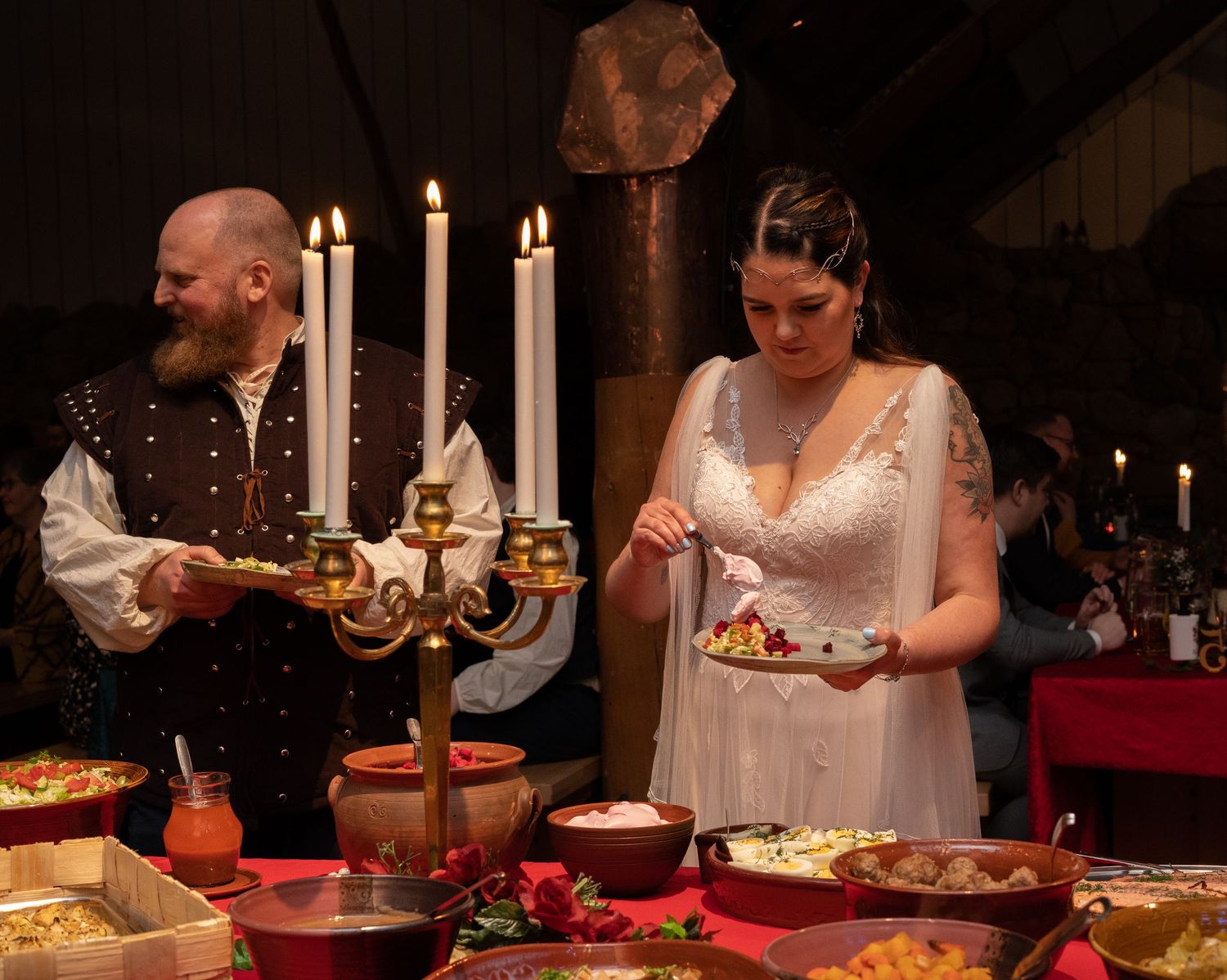 Medieval-themed wedding couple serving food at a festive banquet table with candelabra and colorful dishes.