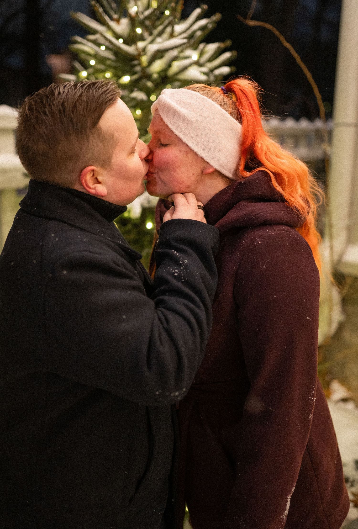 Couple sharing a romantic kiss outdoors near a snow-dusted Christmas tree, woman with orange hair wearing a pink headband.