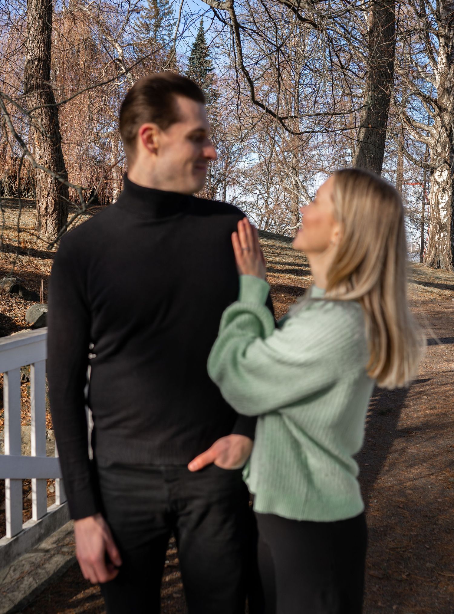 A young couple shares a tender moment outdoors in a park, woman in green sweater gently touching mans chest.