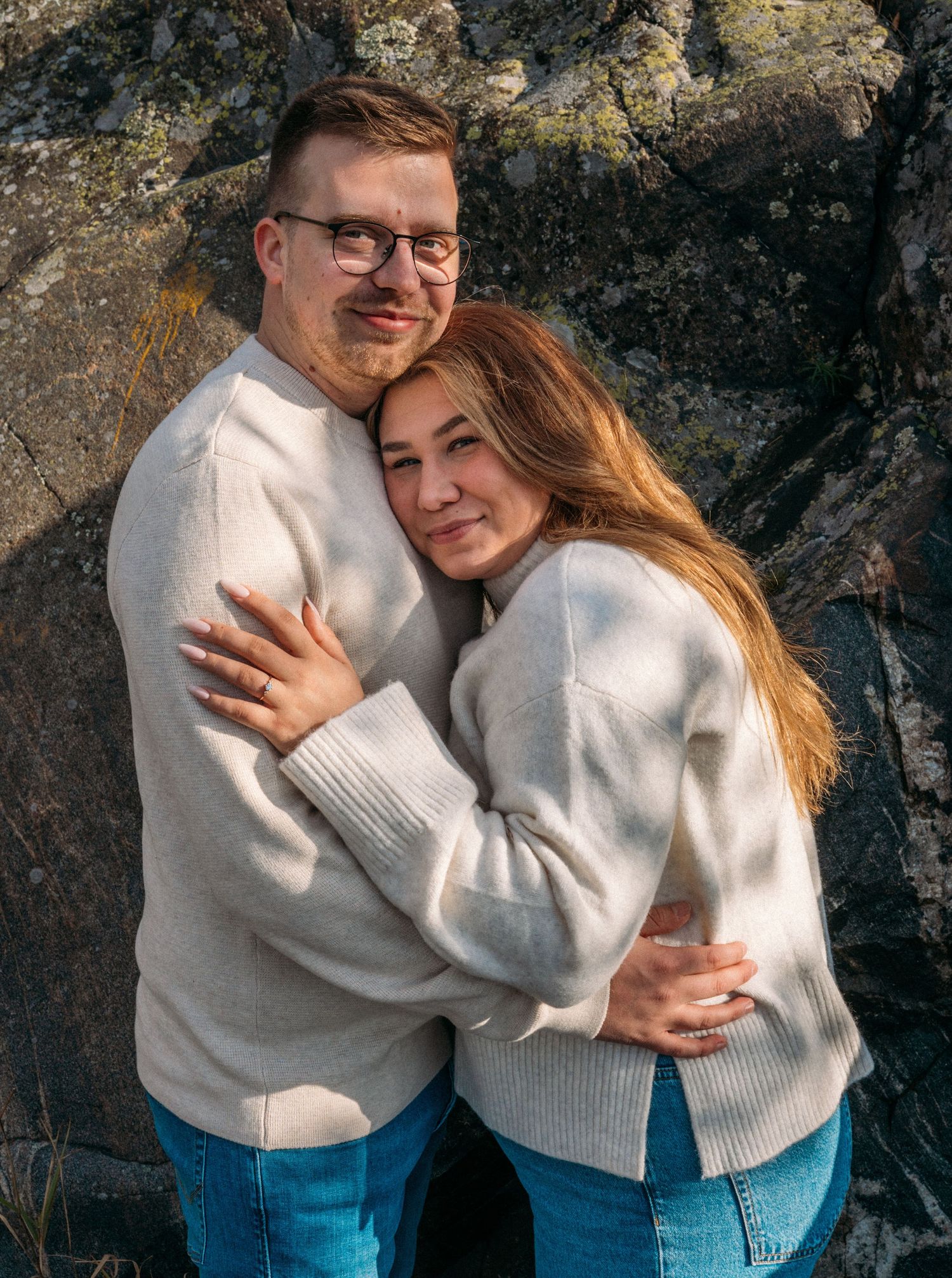 A couple in matching cream sweaters and jeans embrace warmly against a rocky backdrop outdoors.