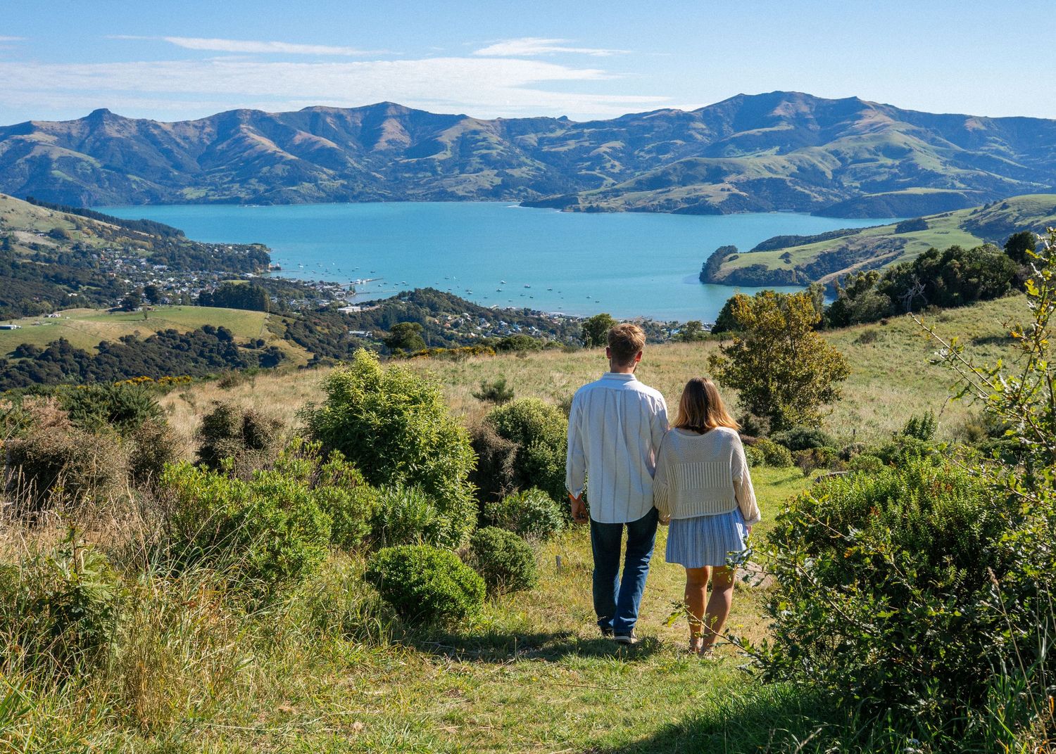 Romantic couple holding hands walking through lush green hills overlooking a stunning blue bay and mountain vista.