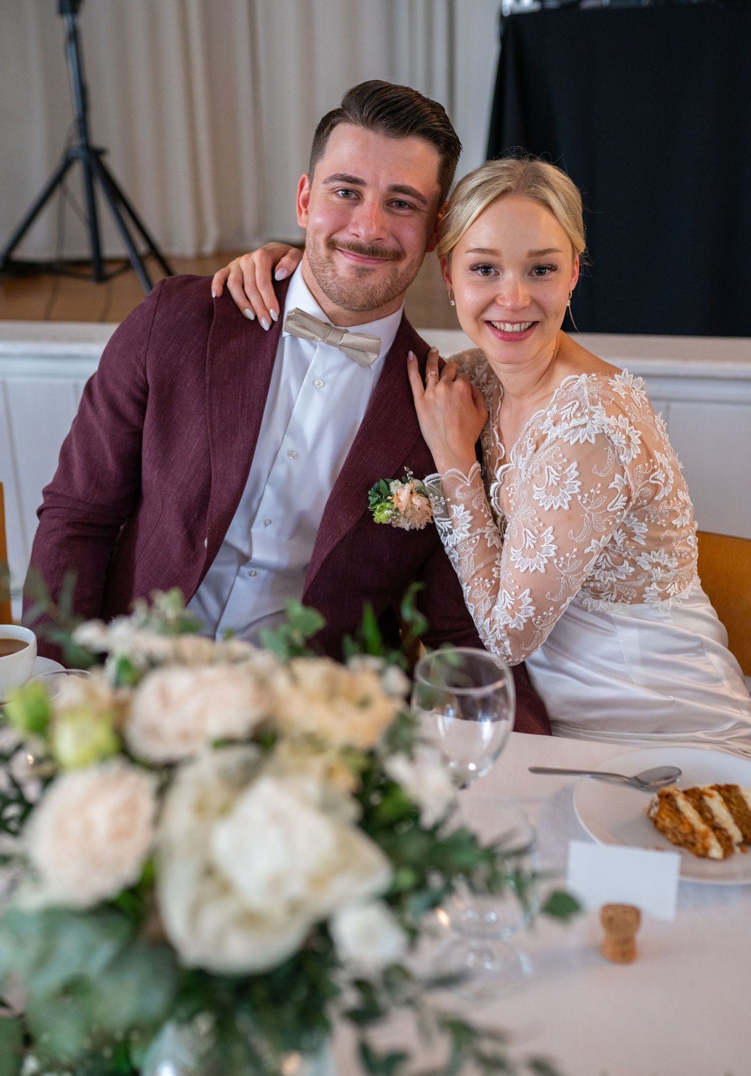 Smiling bride and groom in burgundy suit seated at wedding reception table with white flowers.