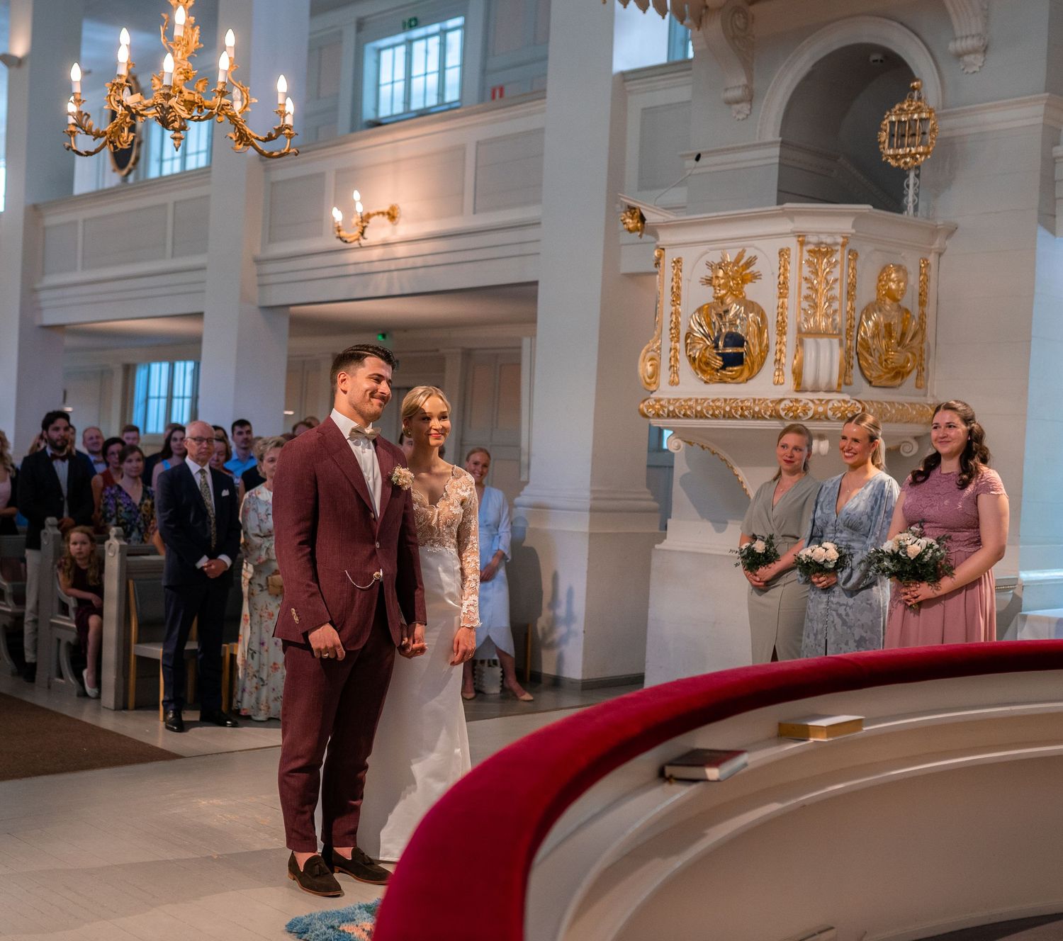 Bride and groom stand smiling at church altar with bridesmaids and chandelier during wedding ceremony.
