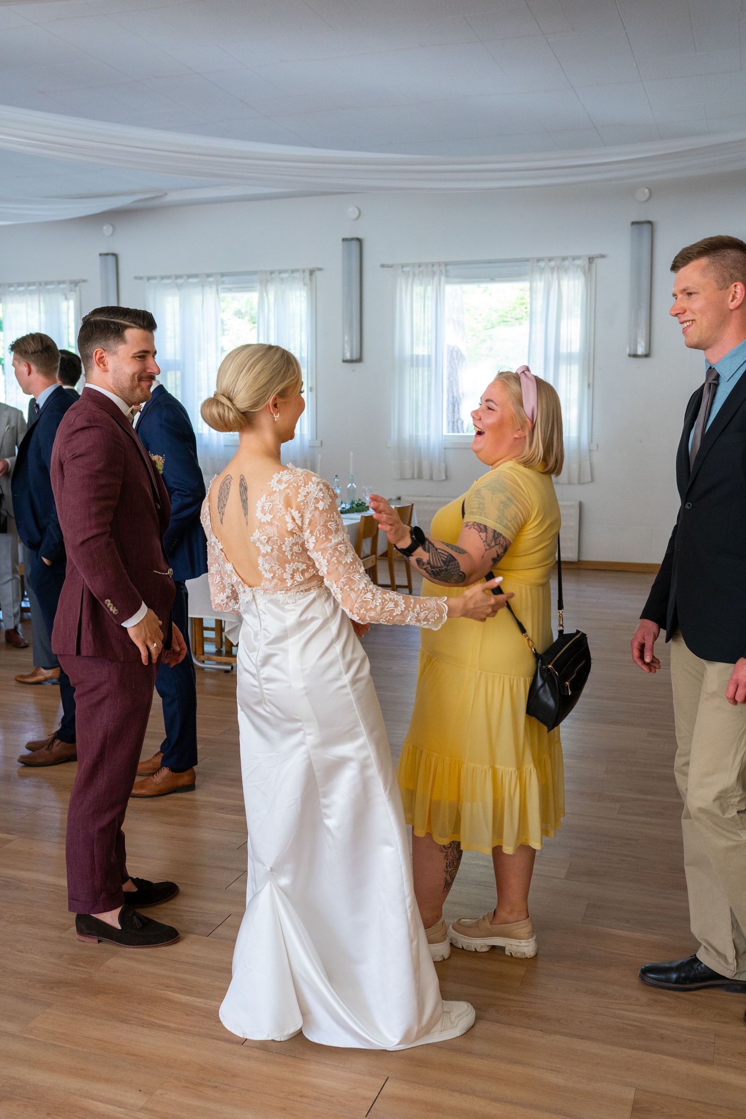 Bride in backless lace gown chats with laughing guest in yellow dress at wedding reception.