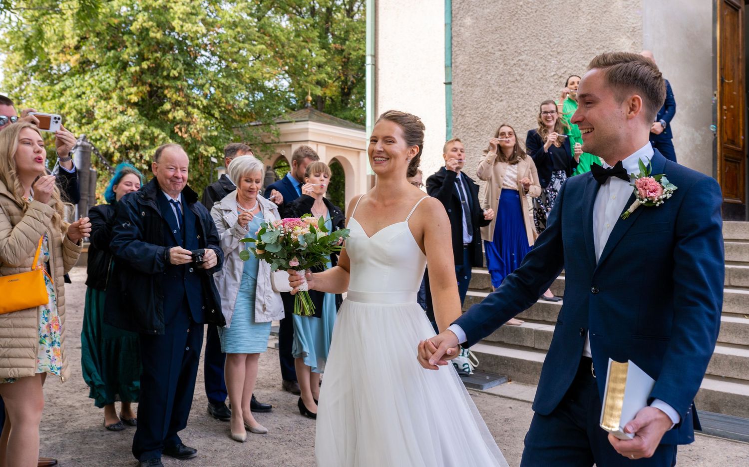 Newlyweds hold hands walking from church as guests celebrate behind them.