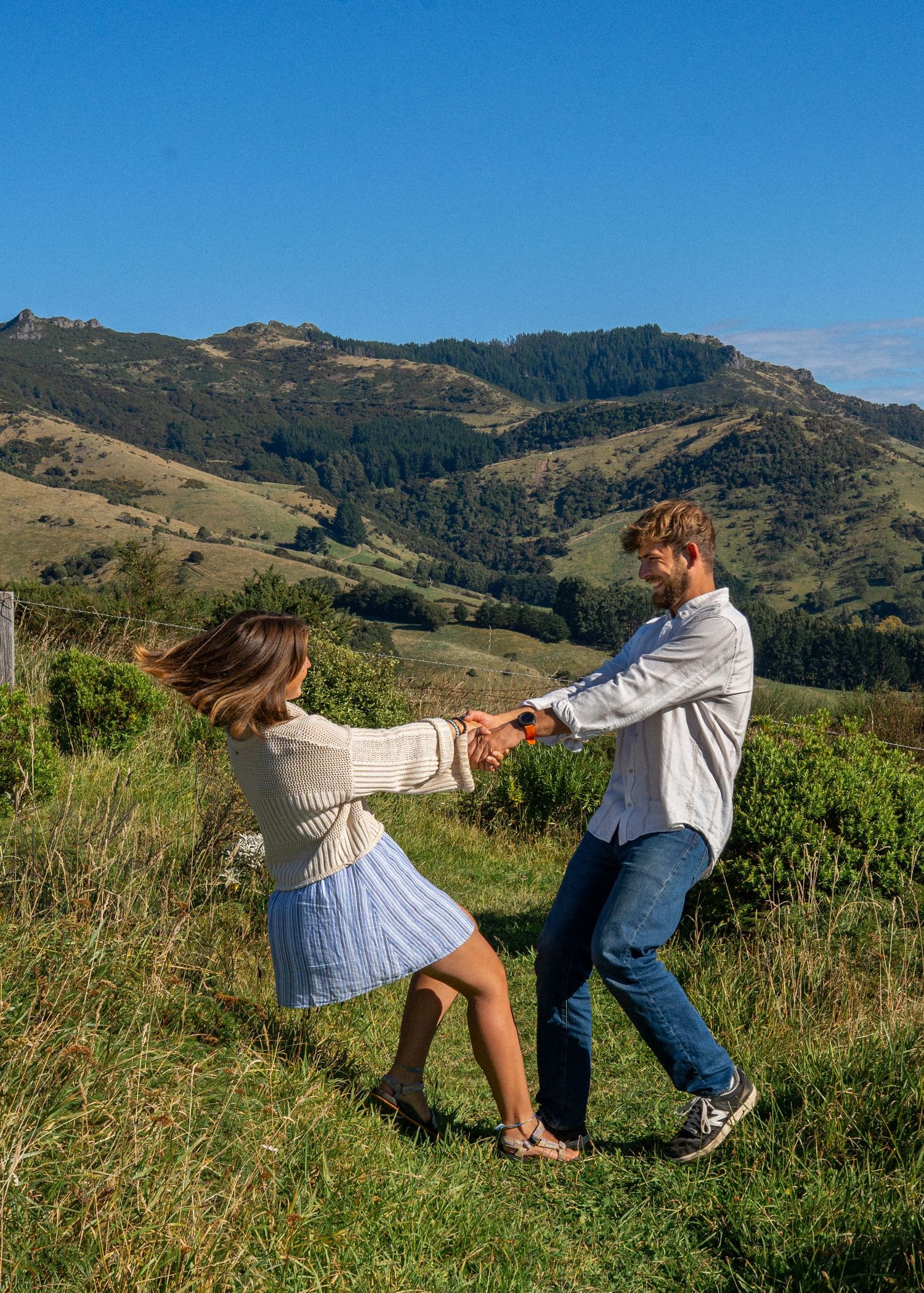 A joyful couple dances and spins together on a green hillside with scenic mountain views under a clear blue sky.
