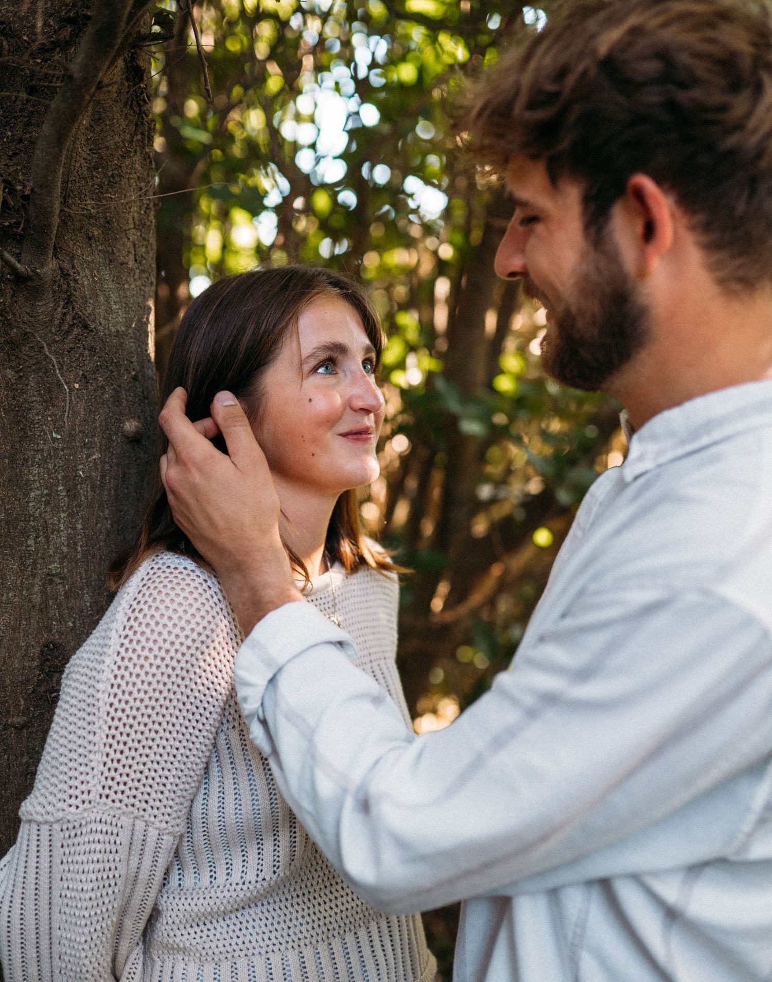 Man tenderly holds woman's face near a tree in a sunlit forest setting.