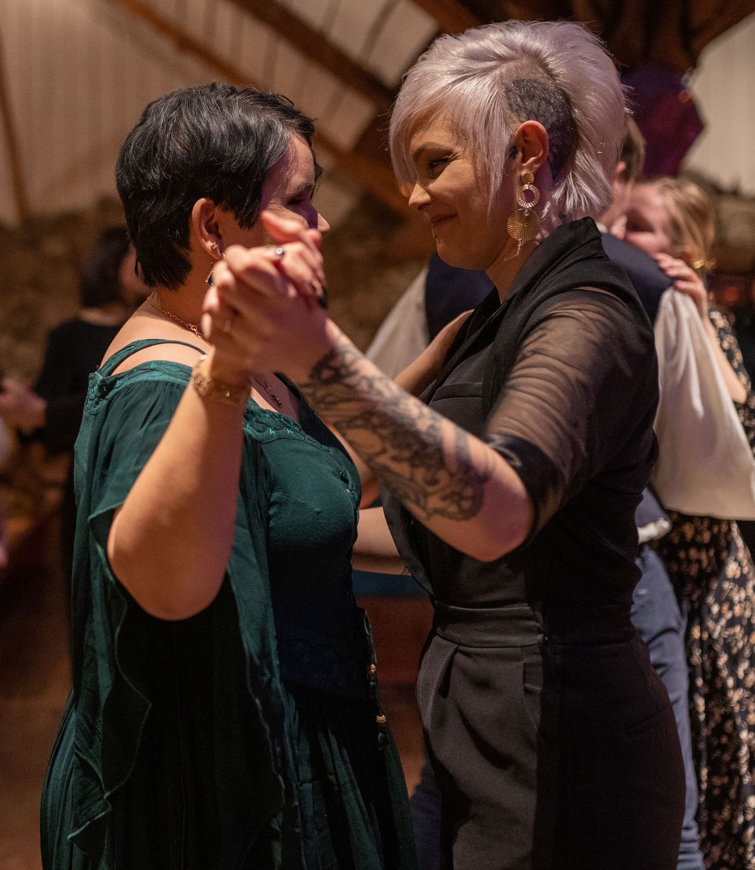Two women dancing together at an indoor event, one in a green dress and one in black with a tattoo sleeve.