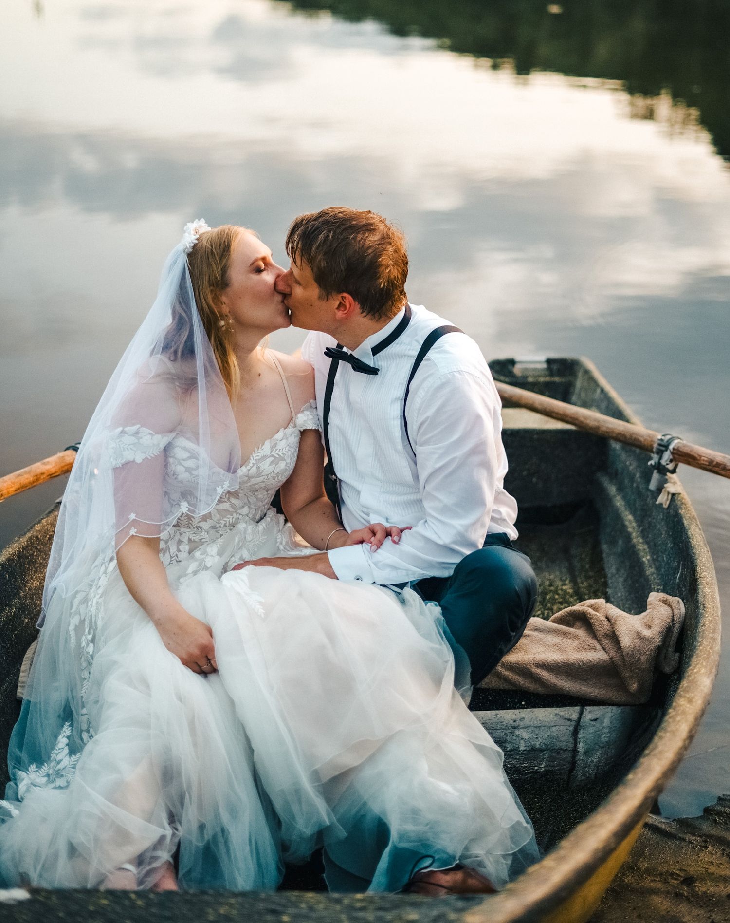 Newlyweds share a tender kiss while sitting in a rustic wooden rowboat on a calm reflective lake.