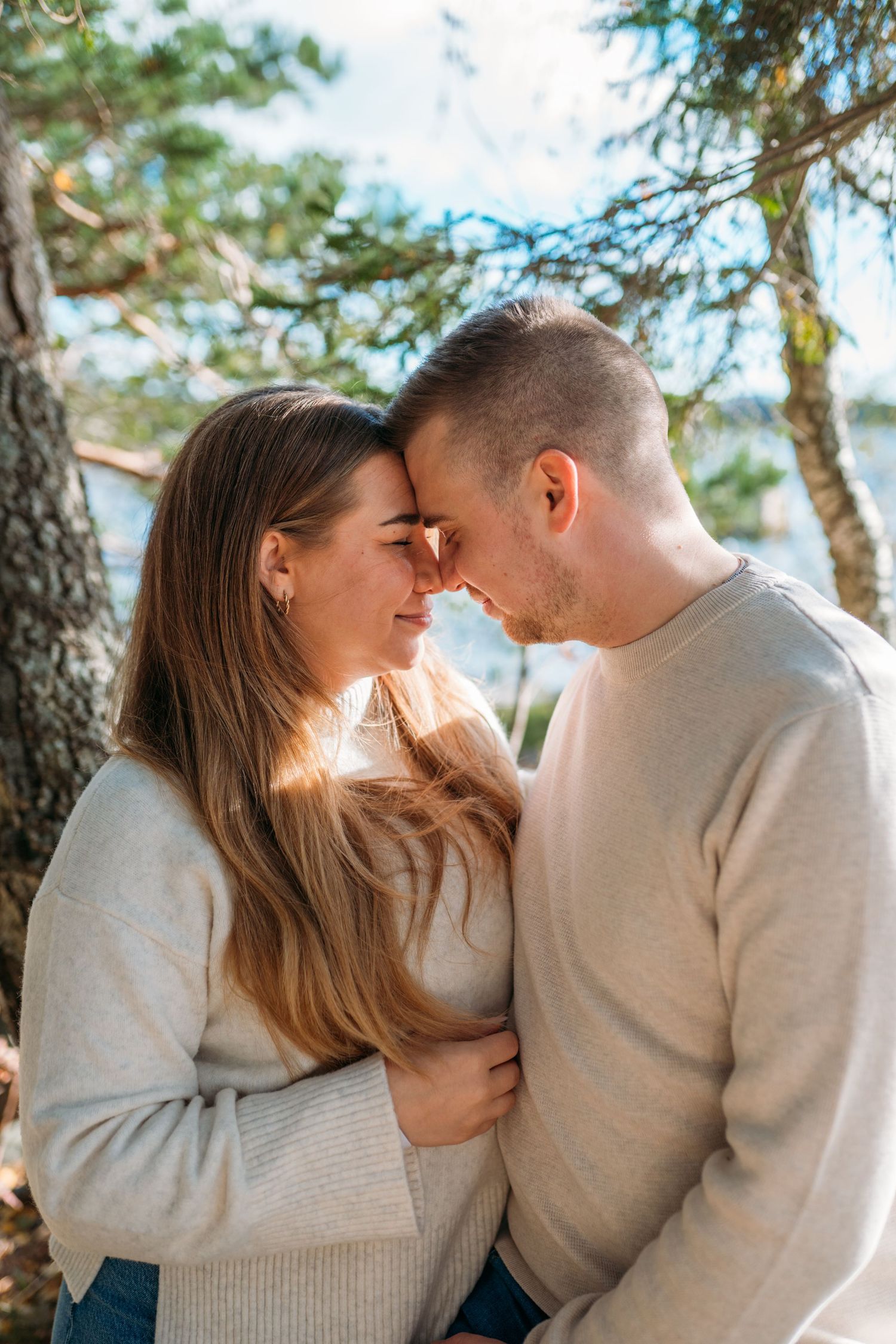 A romantic couple touching foreheads and smiling outdoors among pine trees in matching cream sweaters.