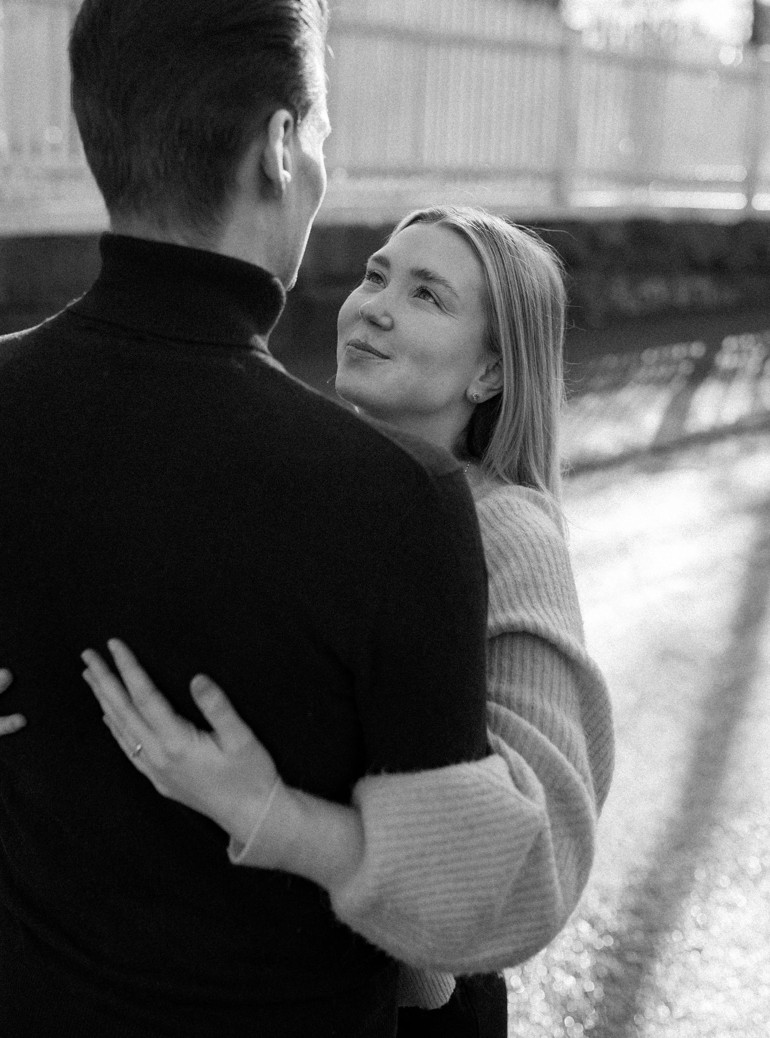 Black and white photo of a couple embracing outdoors, woman smiling up at man in turtleneck sweaters.