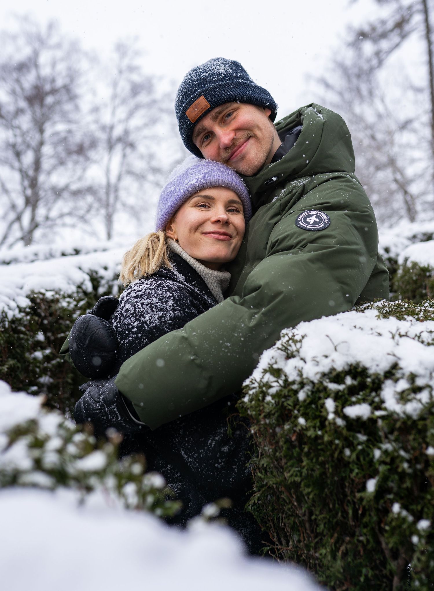Couple in winter hats and jackets embracing outdoors in snowy weather, smiling warmly among snow-covered hedges.