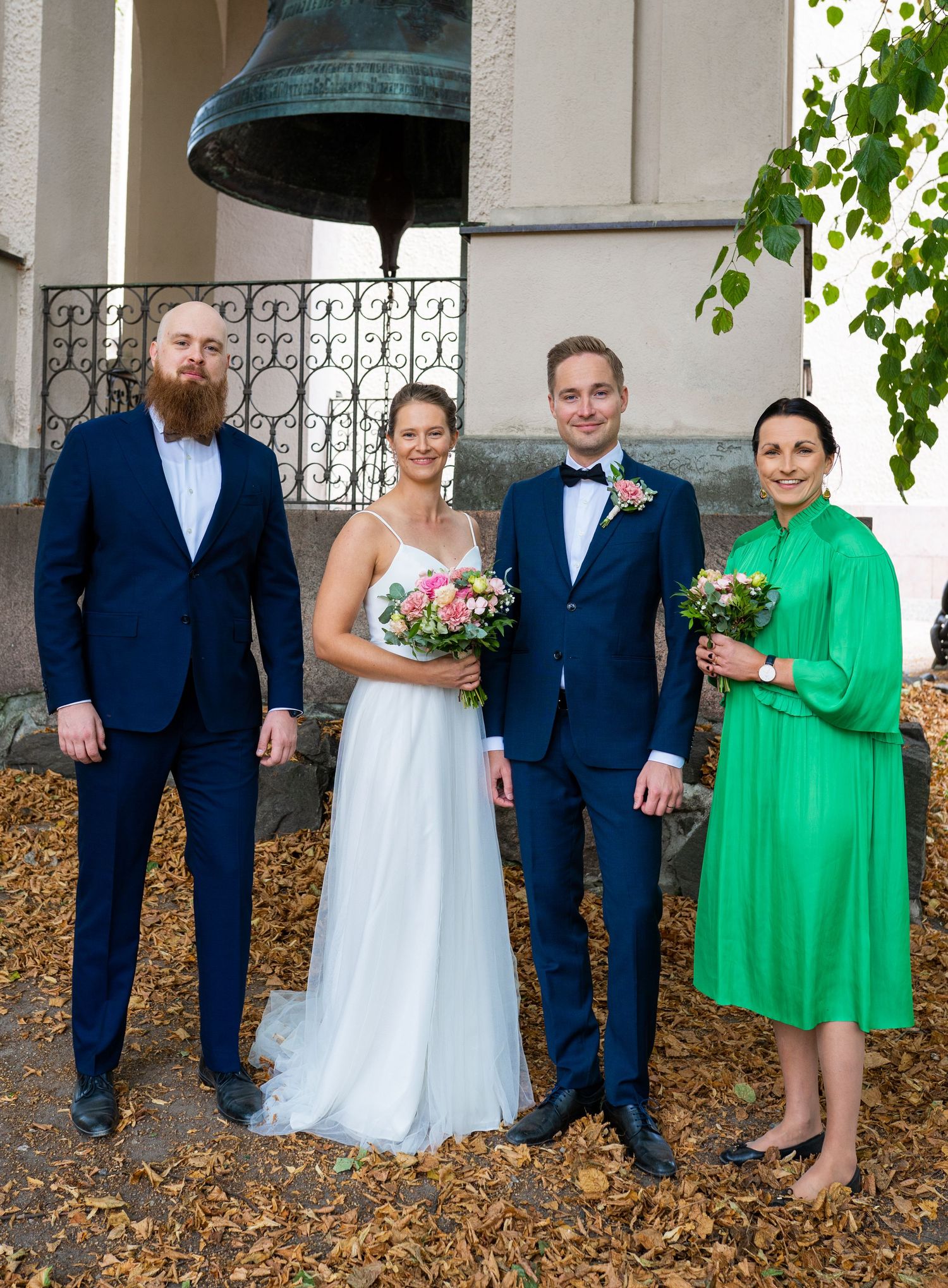 Wedding group of four posing formally near large church bell, couple with witnesses in navy and green attire.