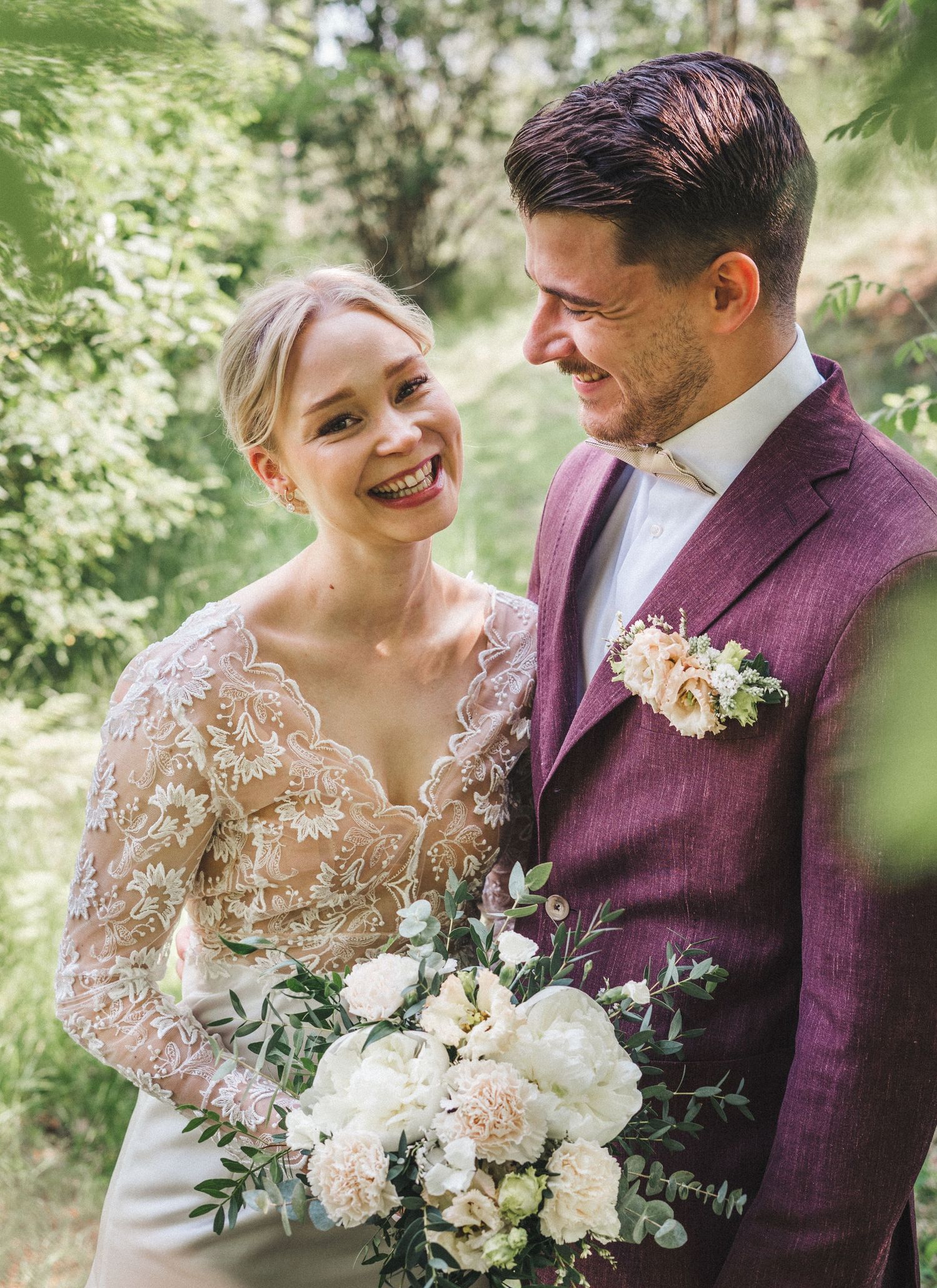 Smiling bride in lace wedding dress gazes at groom in burgundy suit holding white floral bouquet outdoors.