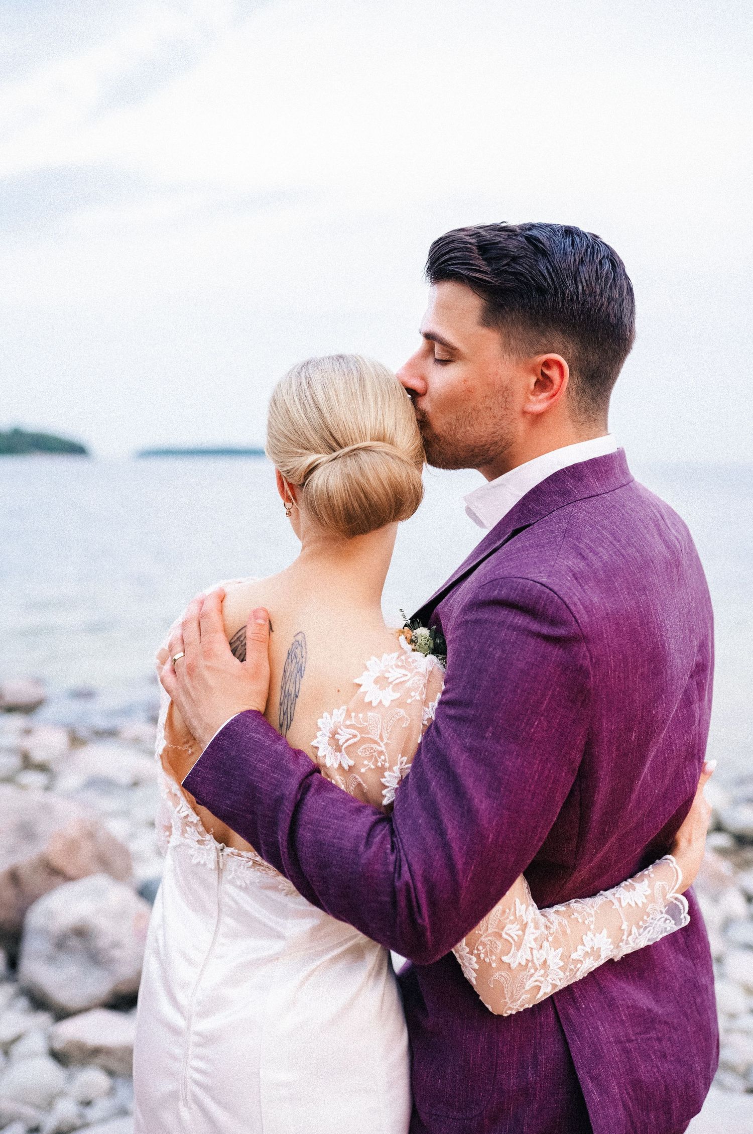 Groom in a purple suit tenderly kissing his bride on the forehead beside a rocky shoreline on their wedding day.