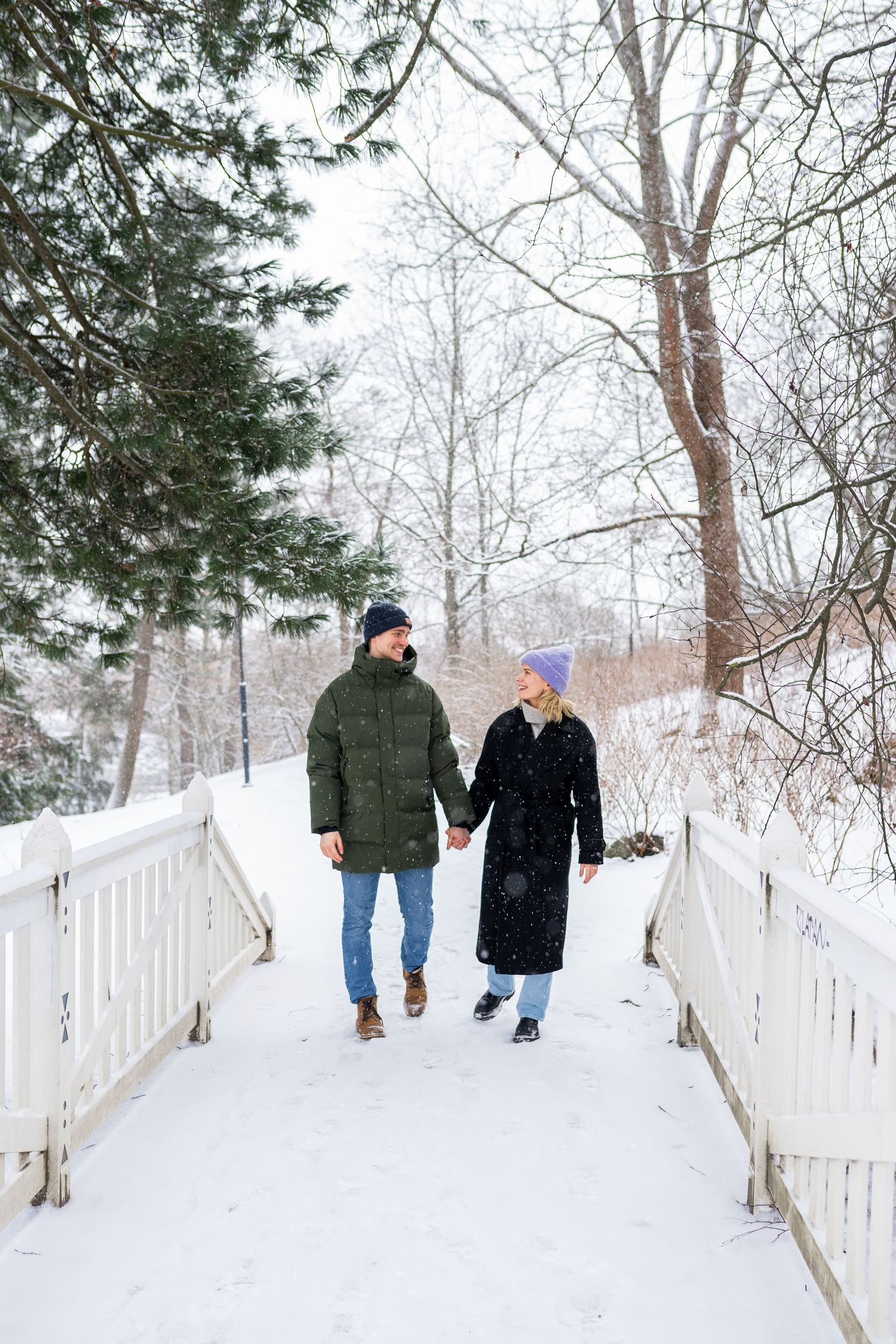 Couple holding hands walking on a snow-covered bridge in a winter park surrounded by snow-dusted trees.