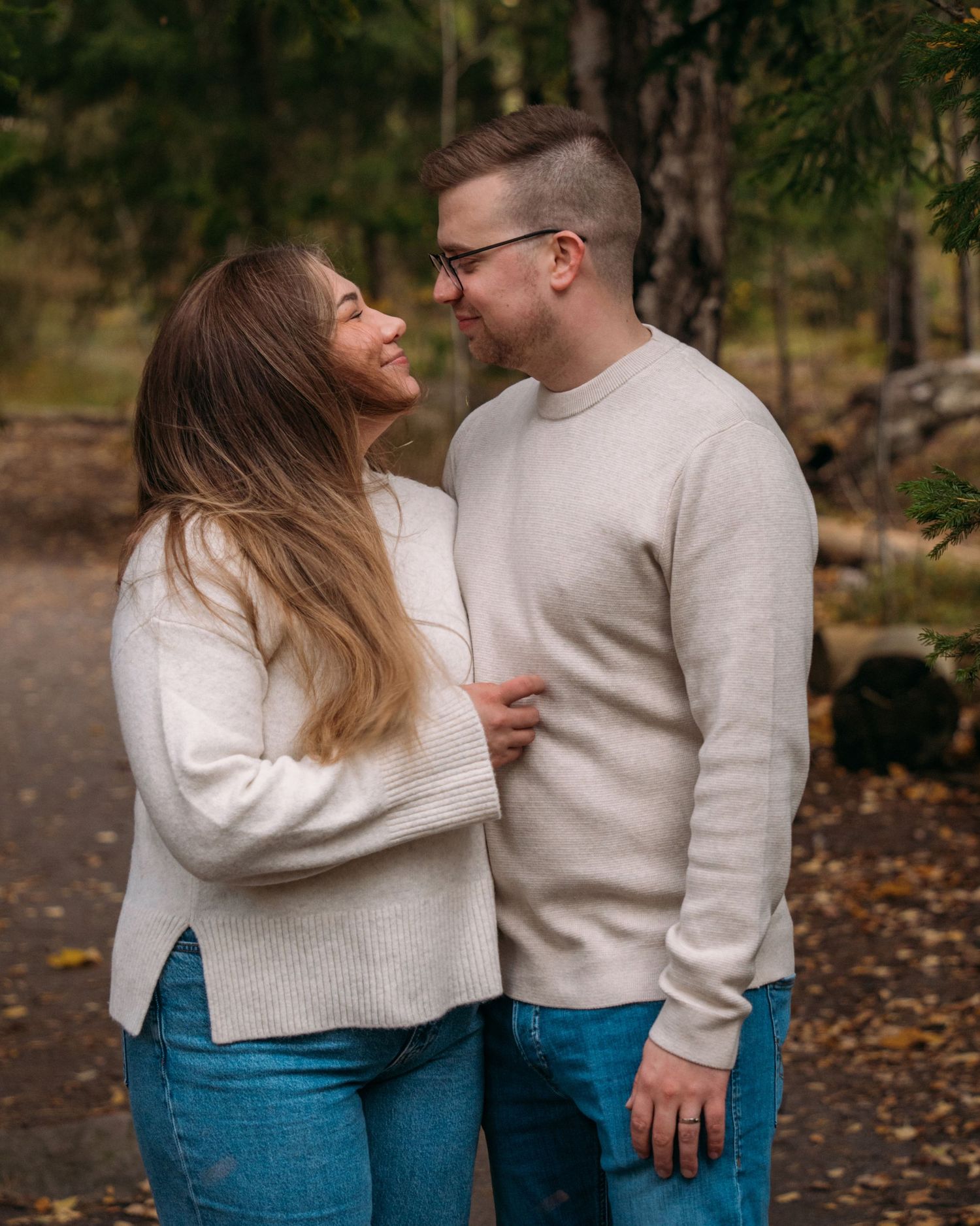 A couple in matching cream sweaters and jeans share a romantic moment on a forest path in autumn.
