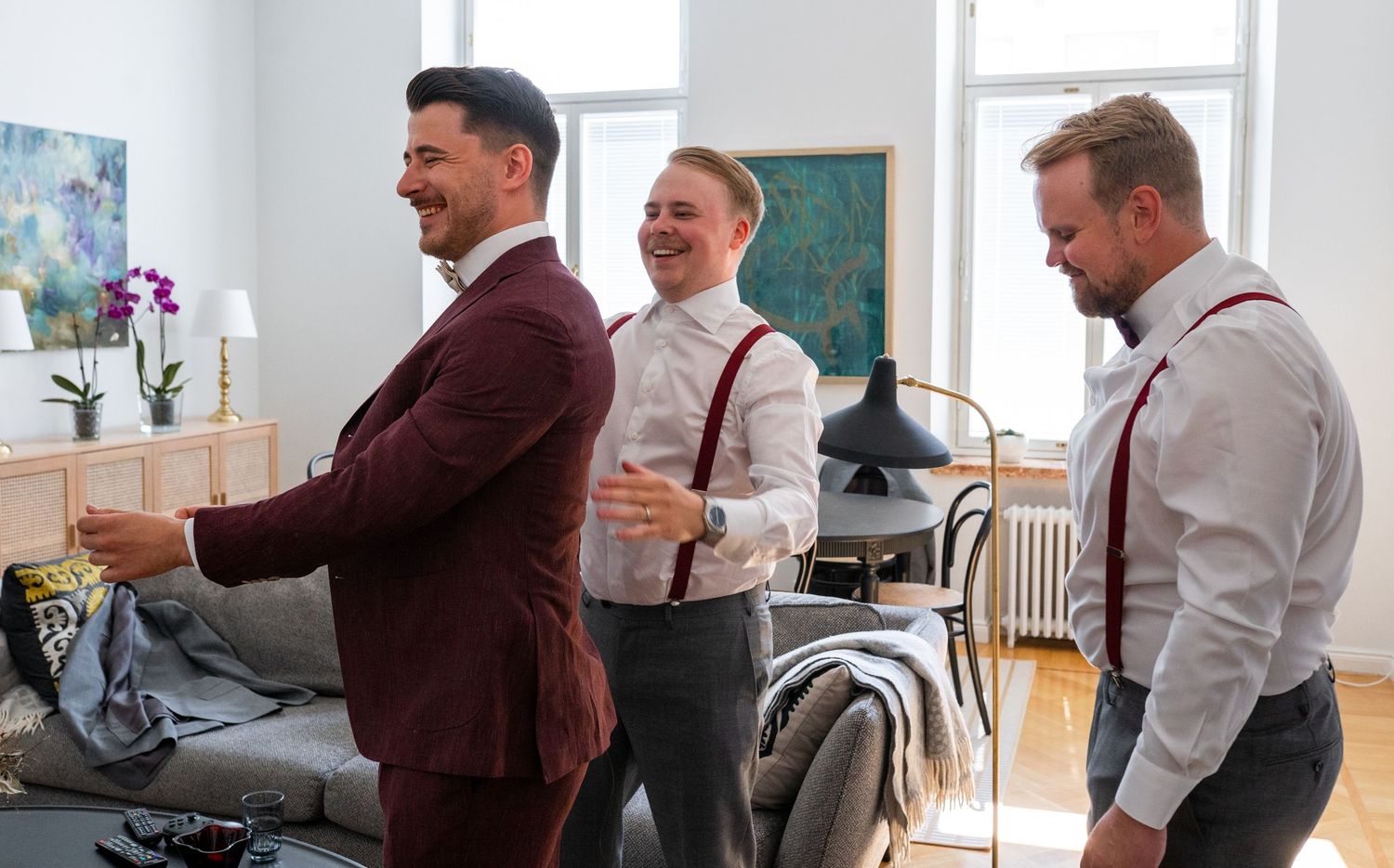 Three men in coordinated burgundy and grey wedding attire sharing a joyful moment while getting dressed.
