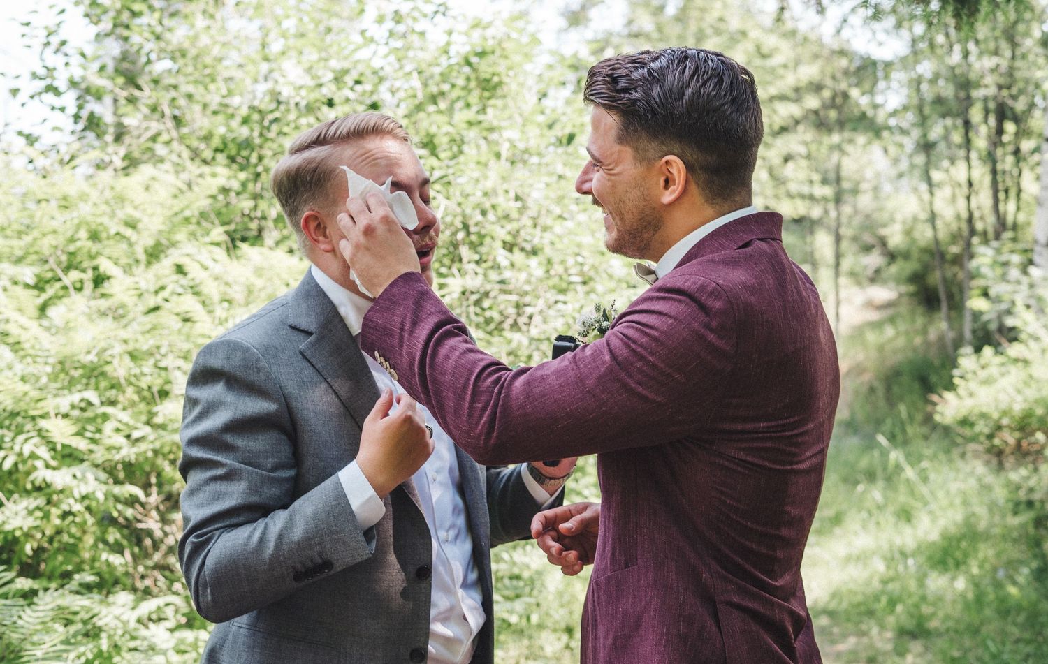 Two men in suits share a playful moment adjusting ties outdoors in a green forest setting.