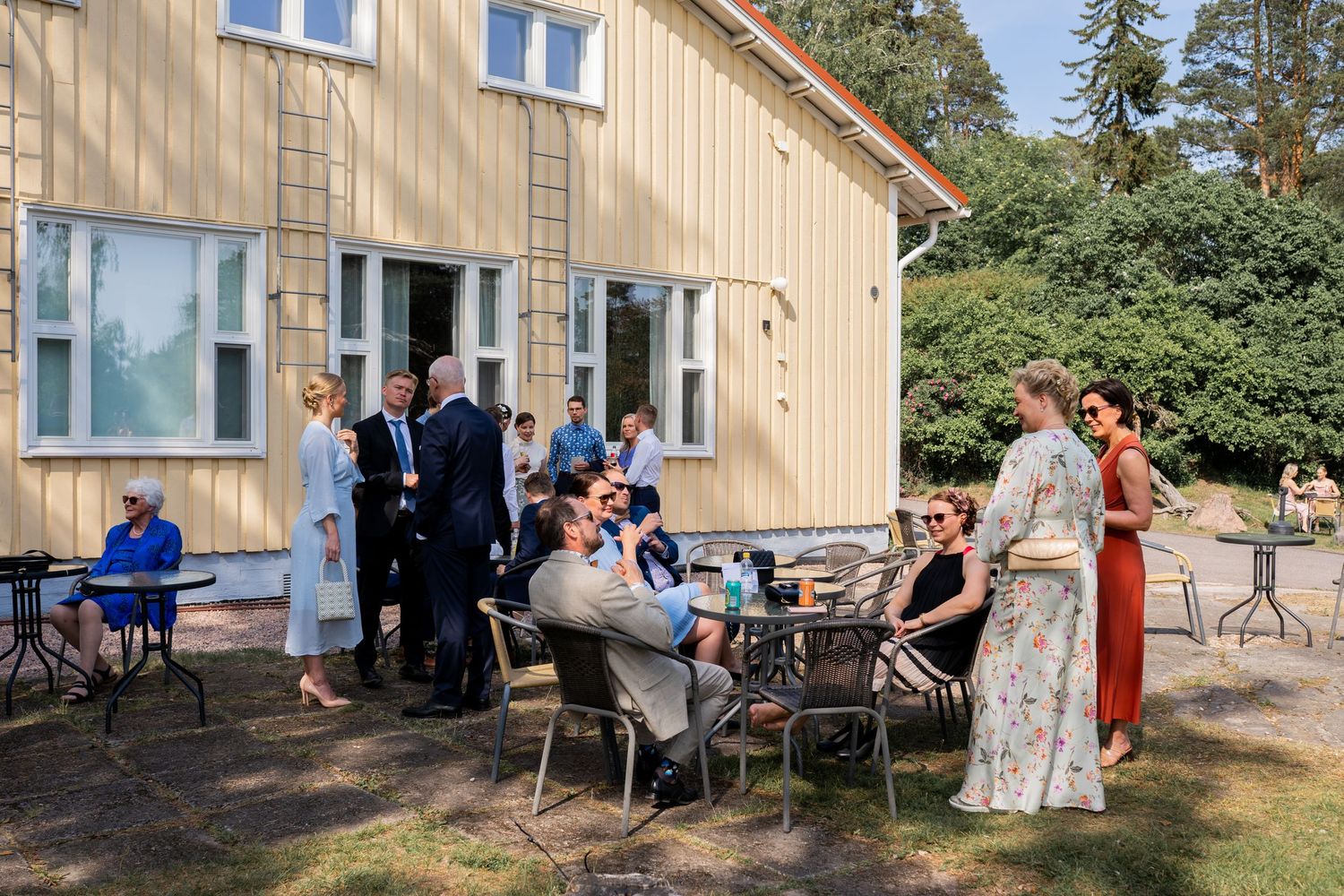 Wedding guests socializing outdoors near a yellow building on a sunny day.