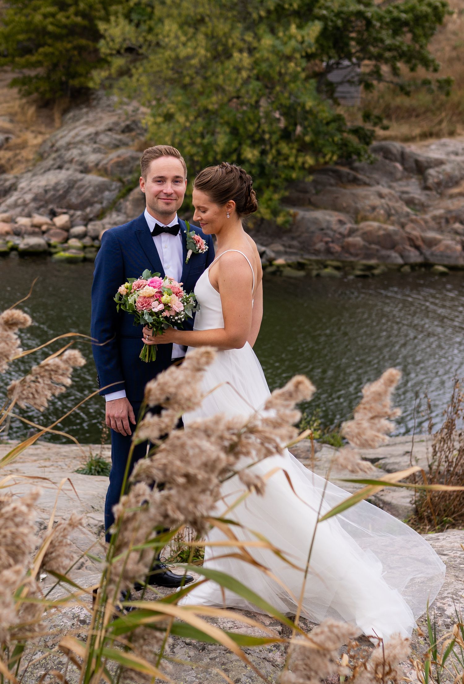 Wedding couple standing on rocks near water, bride holds colorful bouquet, groom in navy tuxedo, tall grass foreground.
